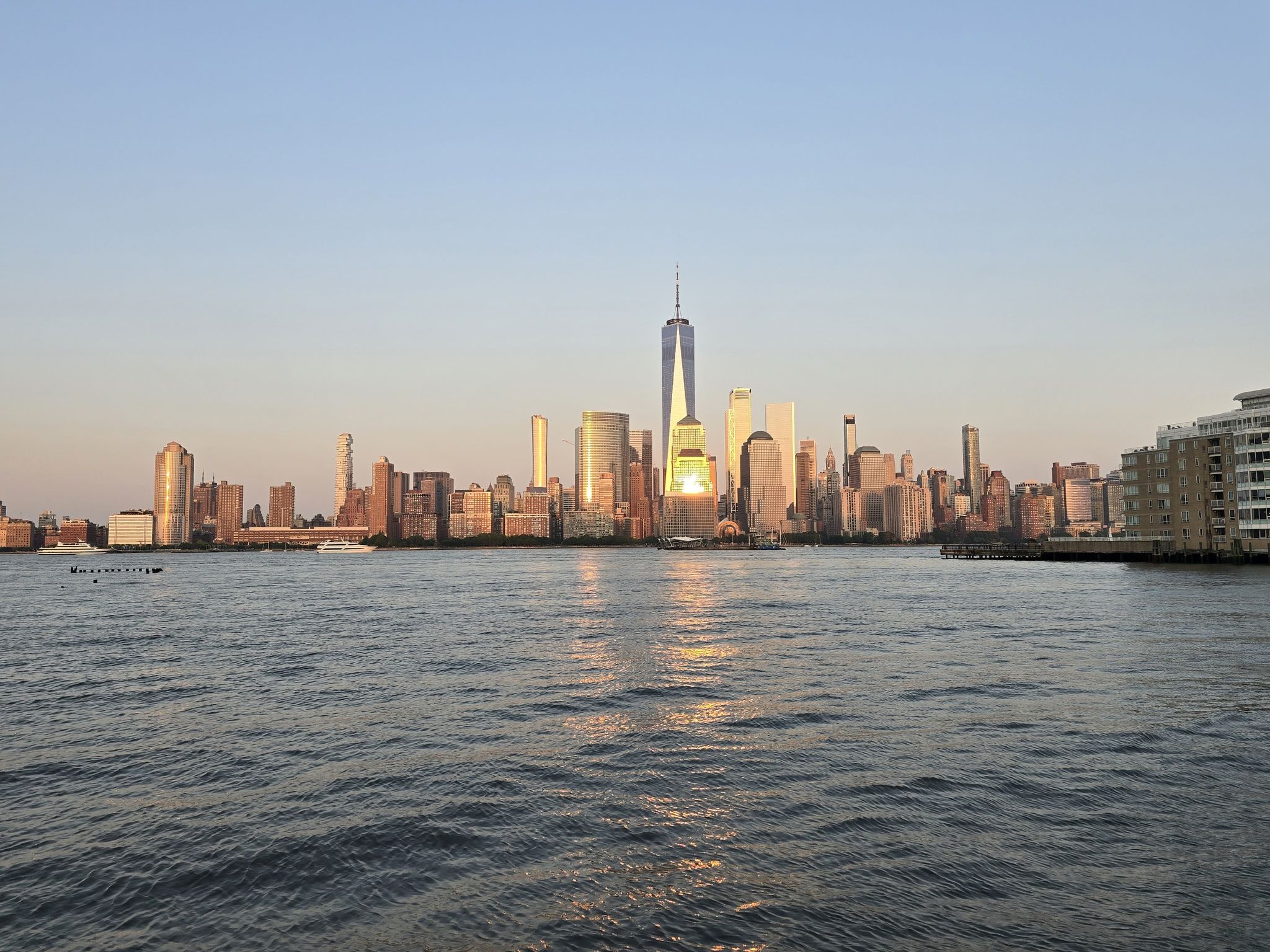 City skyline across water, buildings glowing in golden hour light. - Hoboken Newport Walkway – LeFrak Point Lighthouse loop from Exchange Place