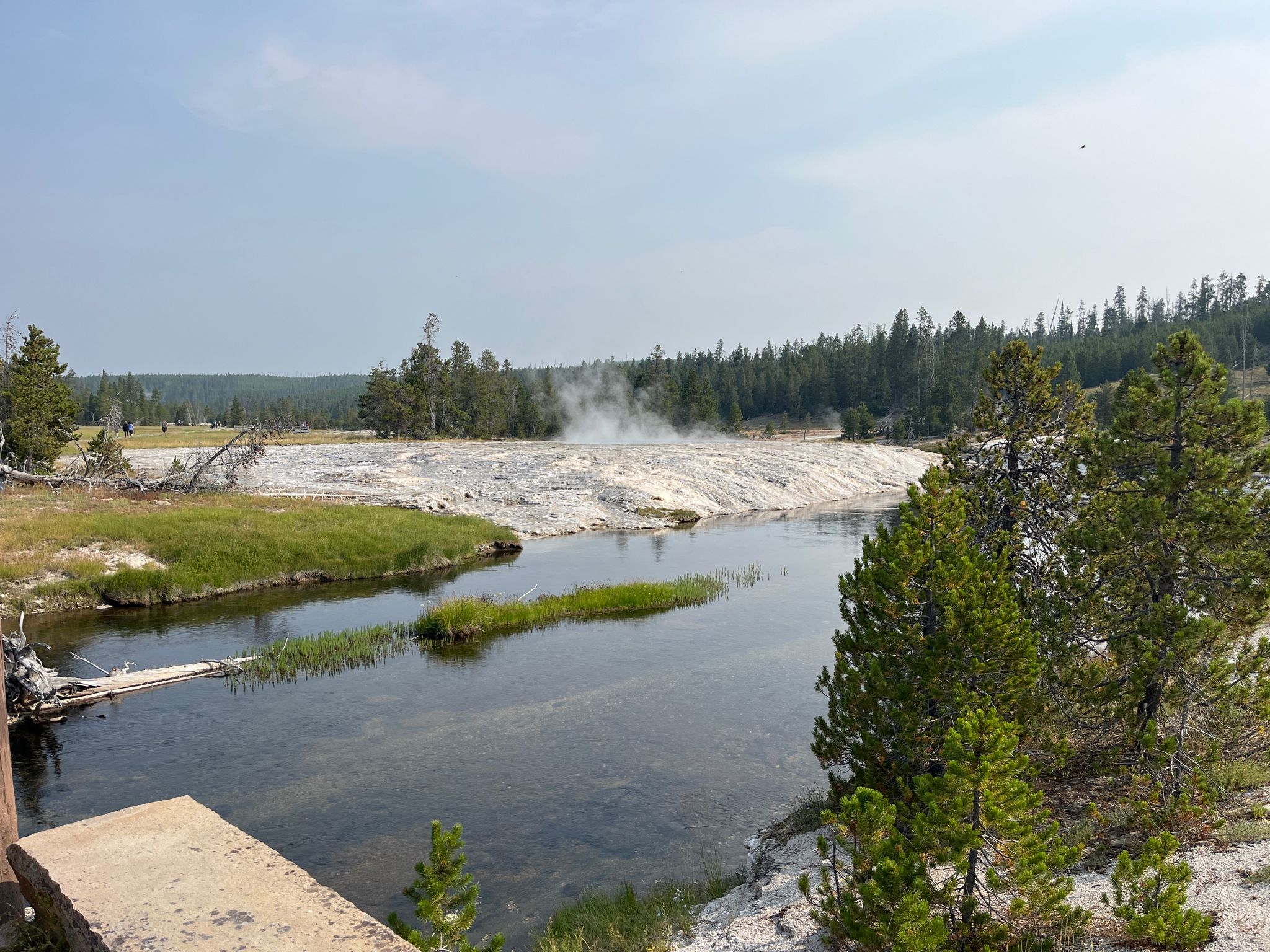 A river flows past a white thermal area with steam rising under a bright sky. - Upper Geyser Basin Trail