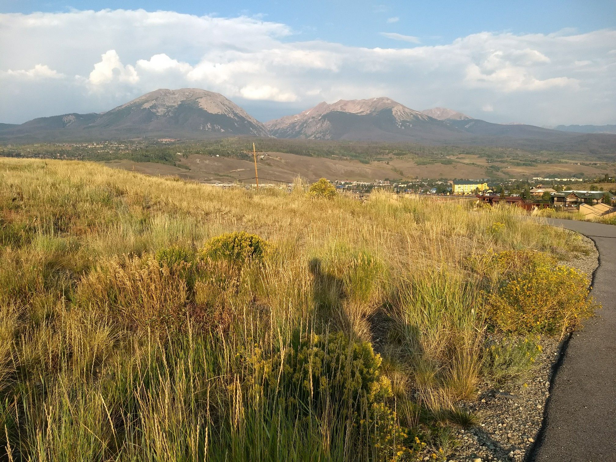Grassy hillside with paved path, distant town, and mountains under clouds. - Silverthorne Blue River Trail – Blue River Trail loop from Silverthorne