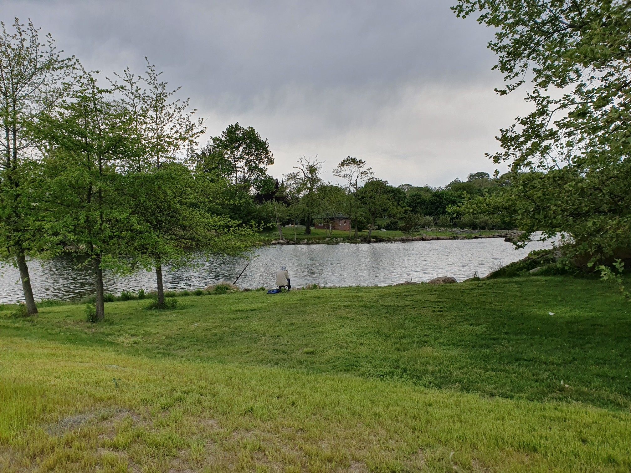 Person fishing by a lake surrounded by trees under a cloudy sky. - Putnam Lake – Putnam Reservoir Dam loop from Cos Cob
