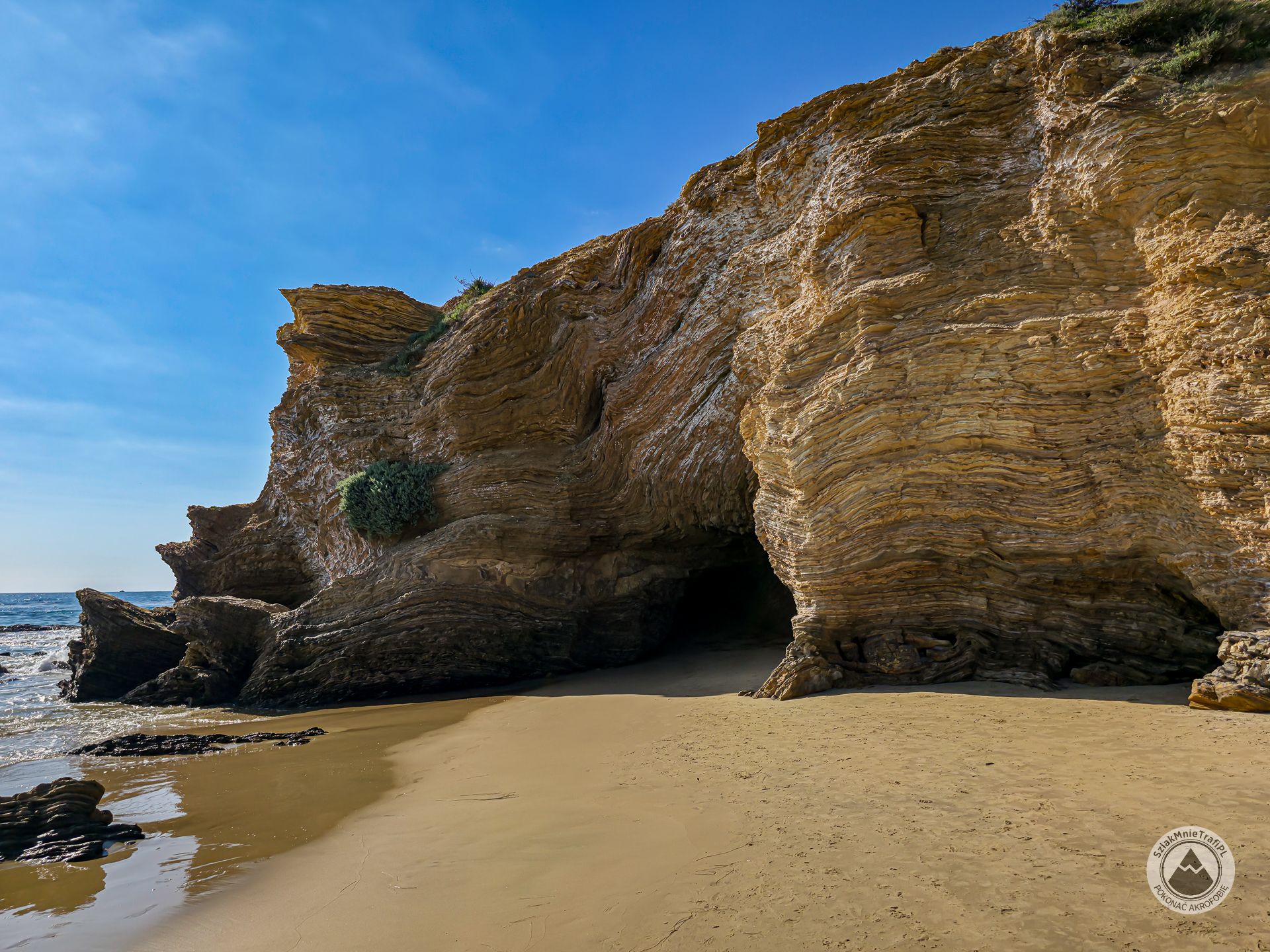 Large layered rock cliff with a cave on a sunny sandy beach. - Blufftop Multiuse Trail