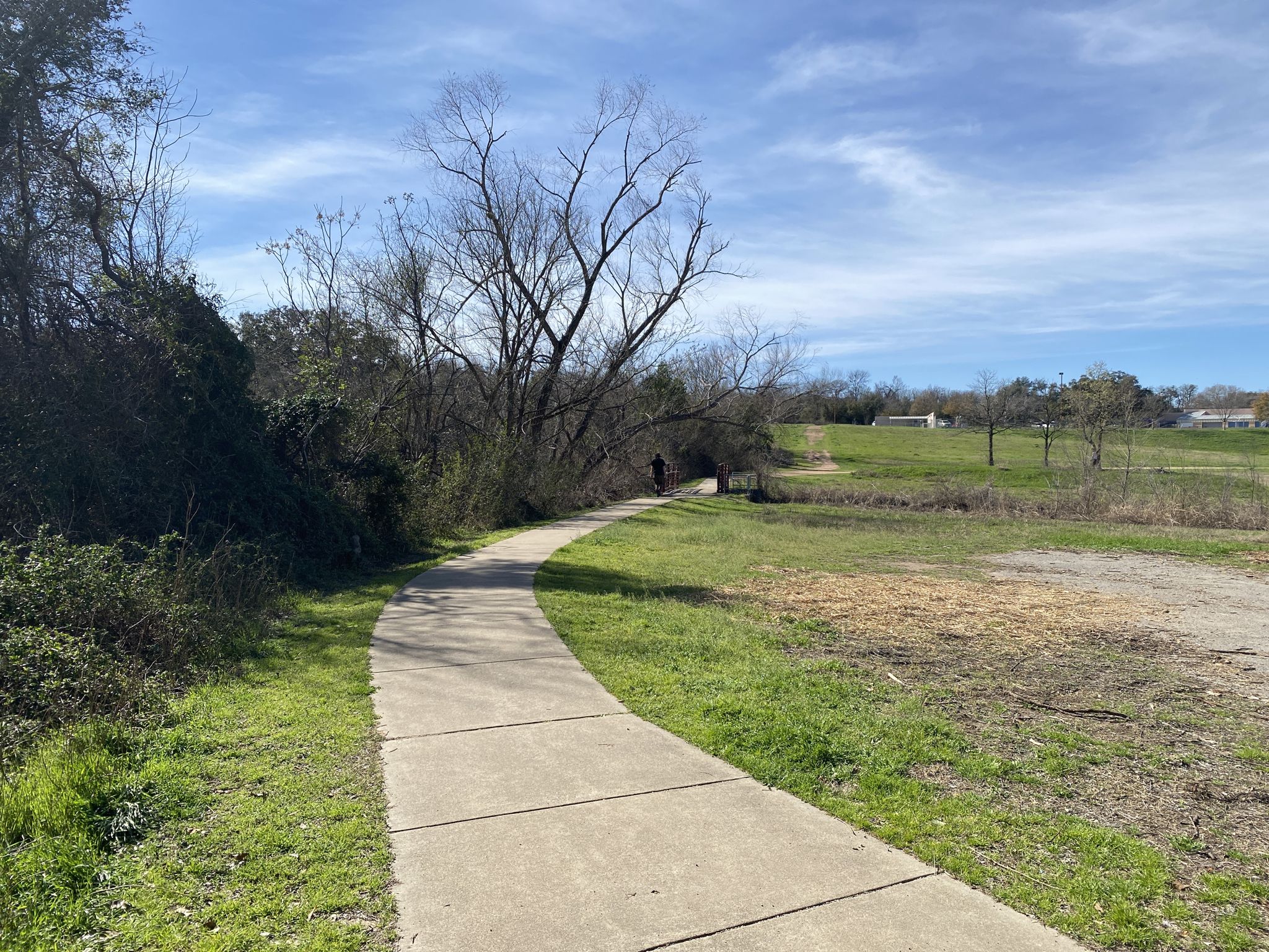 A person walks on a winding paved path through grassy fields under a sunny sky. - Mabel Davis District Park Trail