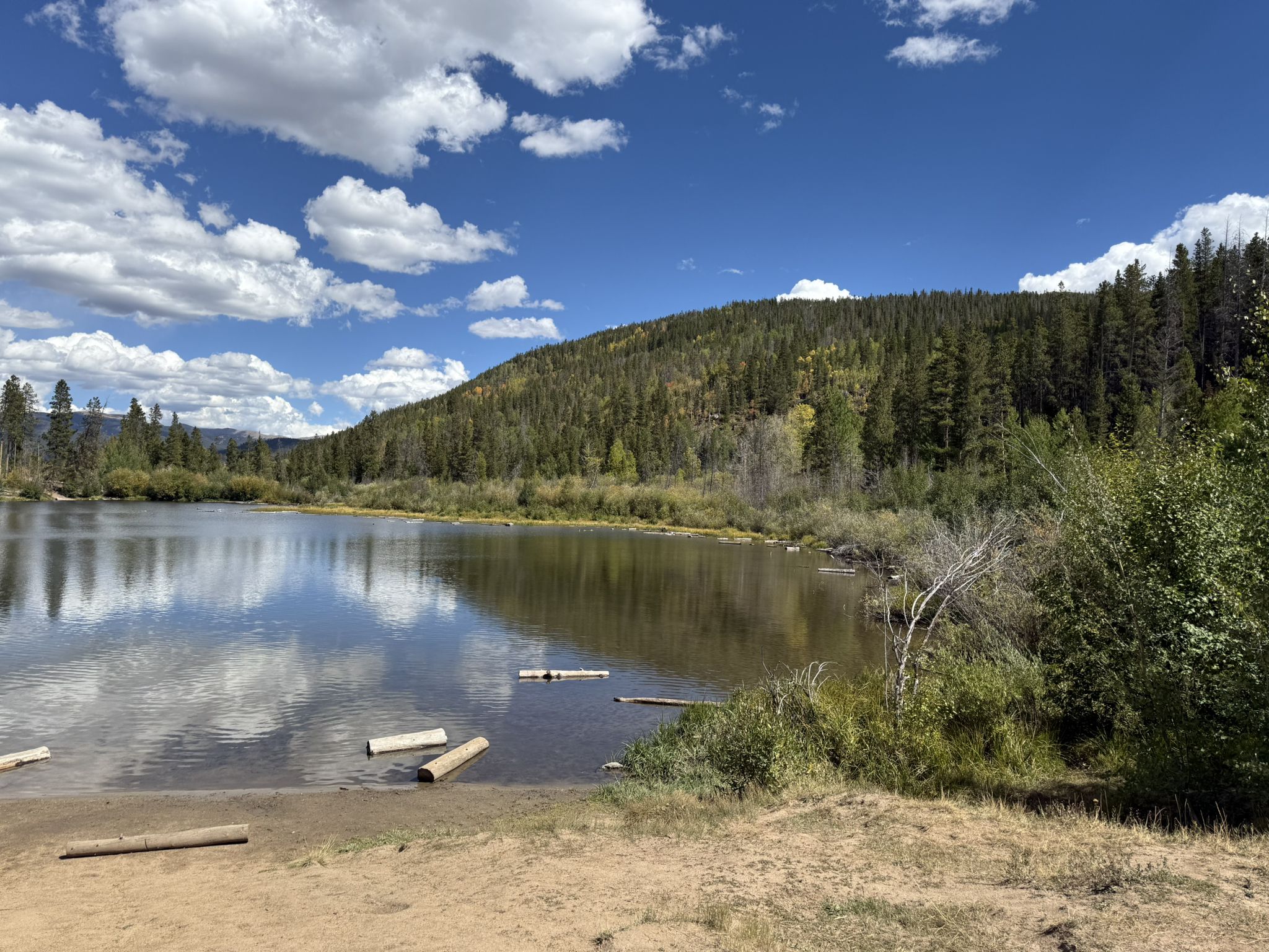 Calm lake reflecting blue sky and clouds, surrounded by green forest. - Peaks Trail and Blue River Recpath Loop