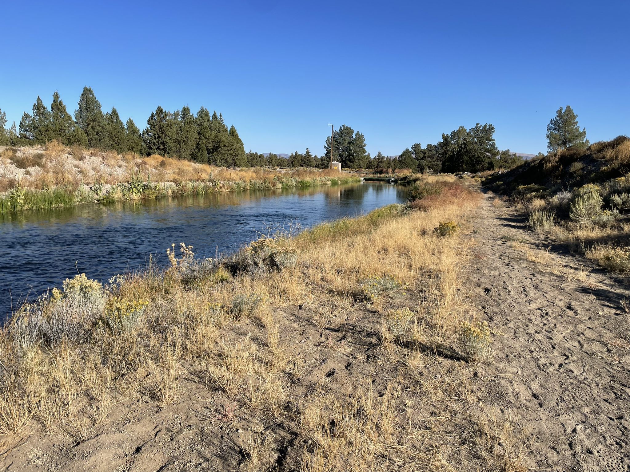 A clear river flows beside a dry dirt path under a bright blue sky. - Hiking loop from Tumulus Trailhead