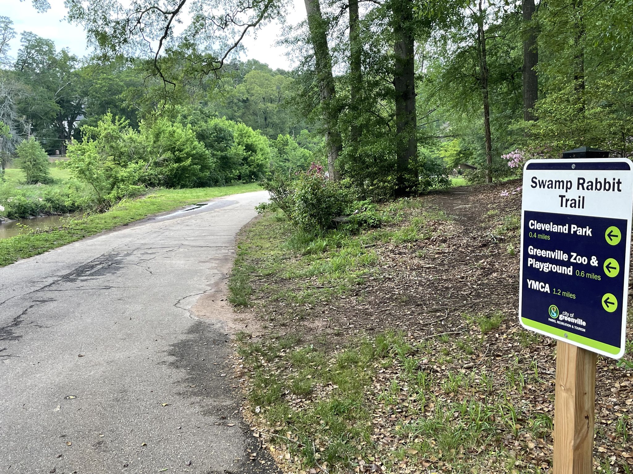 A trail sign stands beside a paved path winding through lush green trees. - Swamp Rabbit Trail to Legacy Park