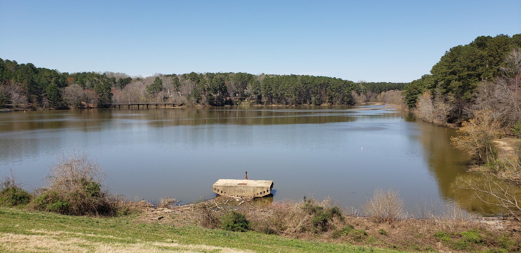Calm lake with forested banks and a concrete structure under a clear sky. - Shelley Lake Trail
