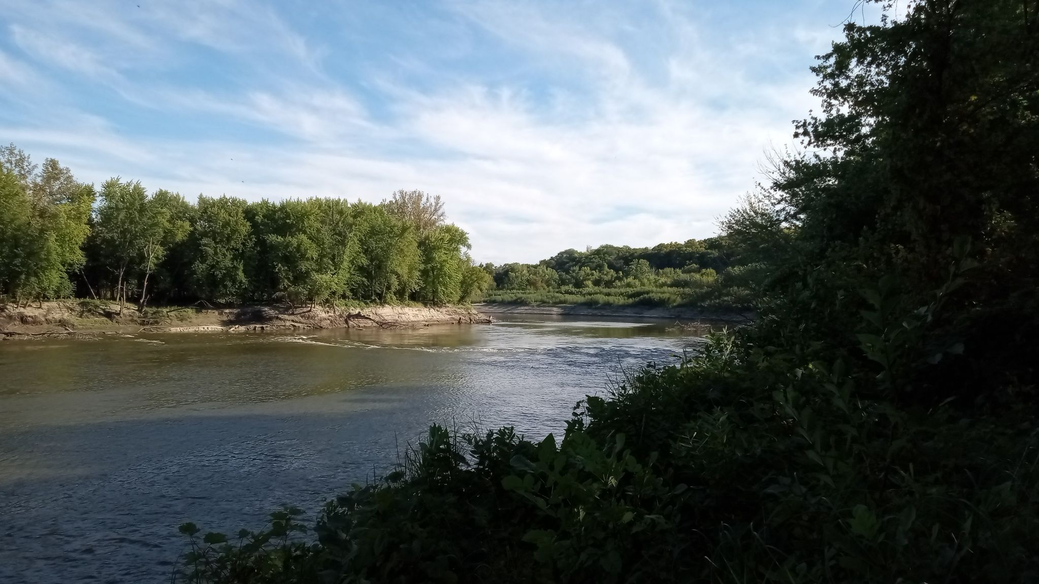 Wide river flowing between lush green tree-lined banks under a bright sky. - Rapids Lake Trail