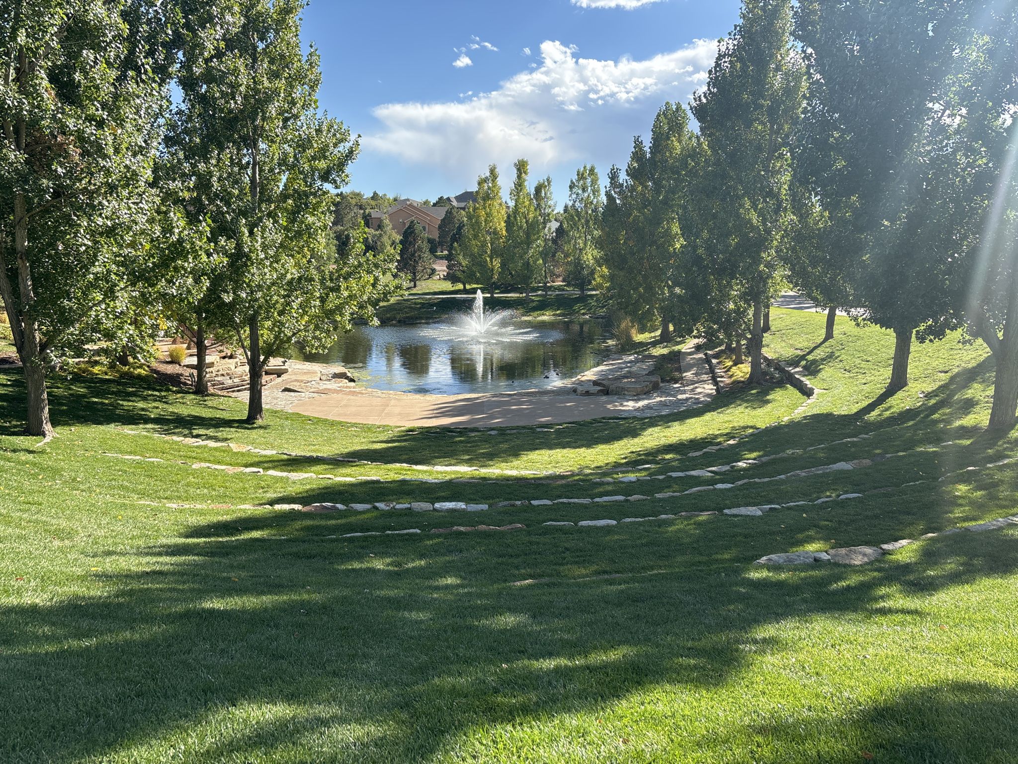 Sunny pond with a fountain, tiered grassy seating, and tall trees. - Mary Kyer Park – Falcon Trail loop from New Life Church