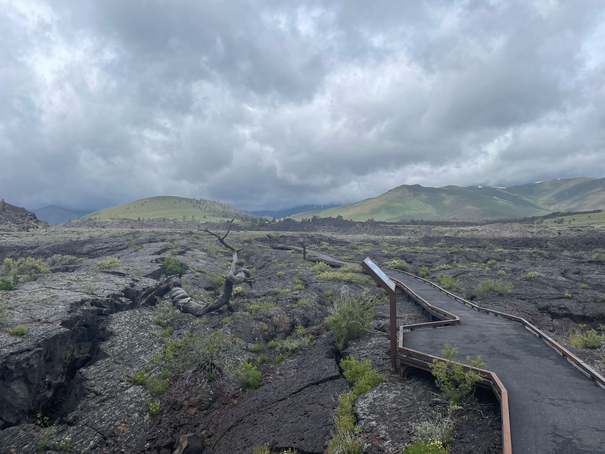 Boardwalk path through cracked volcanic rock and green hills under cloudy sky. - North Crater Flow Trail