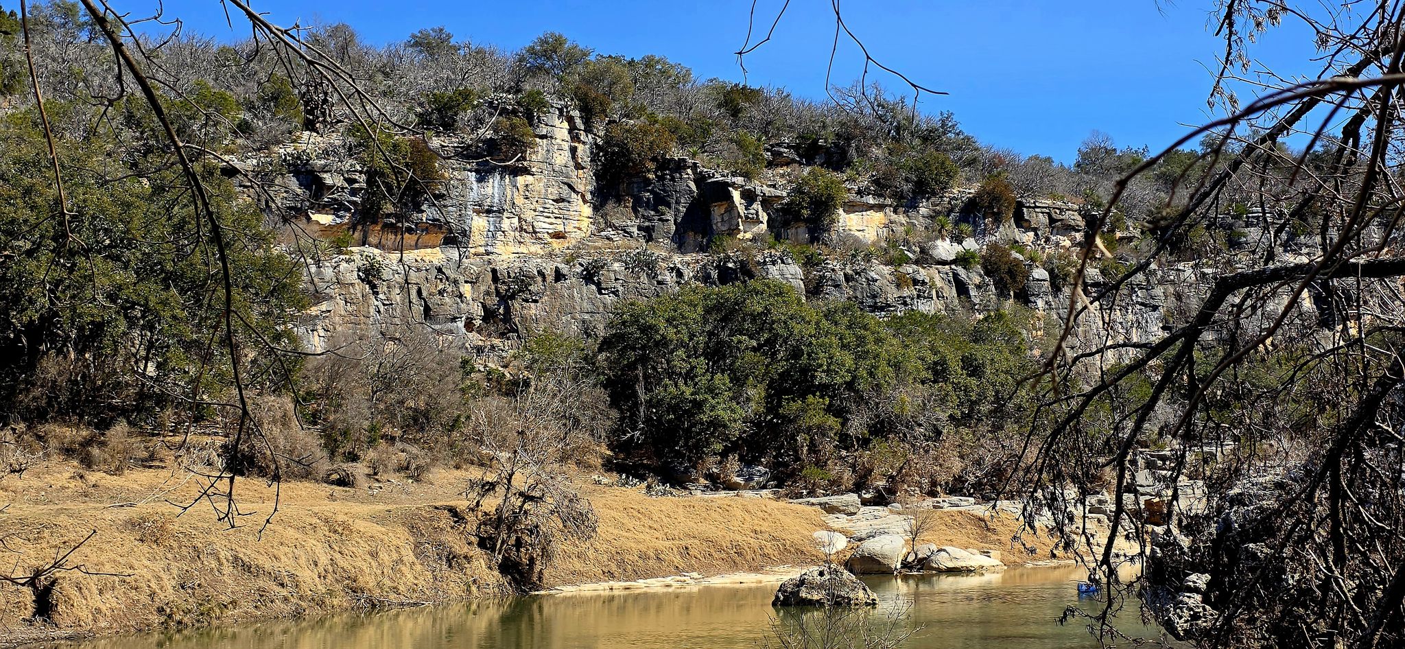 River flowing past rocky cliffs and green trees under a clear sky. - Gorman Falls Trail
