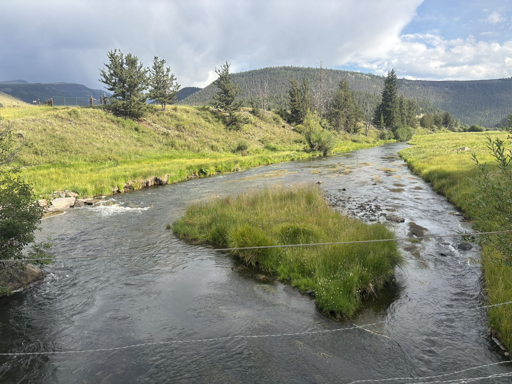 A river splits around a grassy island, surrounded by green hills under a cloudy sky. - Colorado Trail Segment 19 - 21 Detour (La Garita Wilderness Bypass)