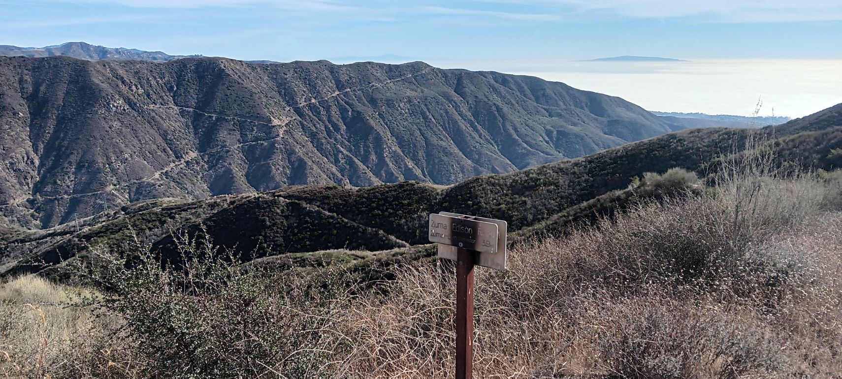 Rugged mountains with trails, dry brush, and a signpost under a clear sky. - Zuma Ridge Trail loop