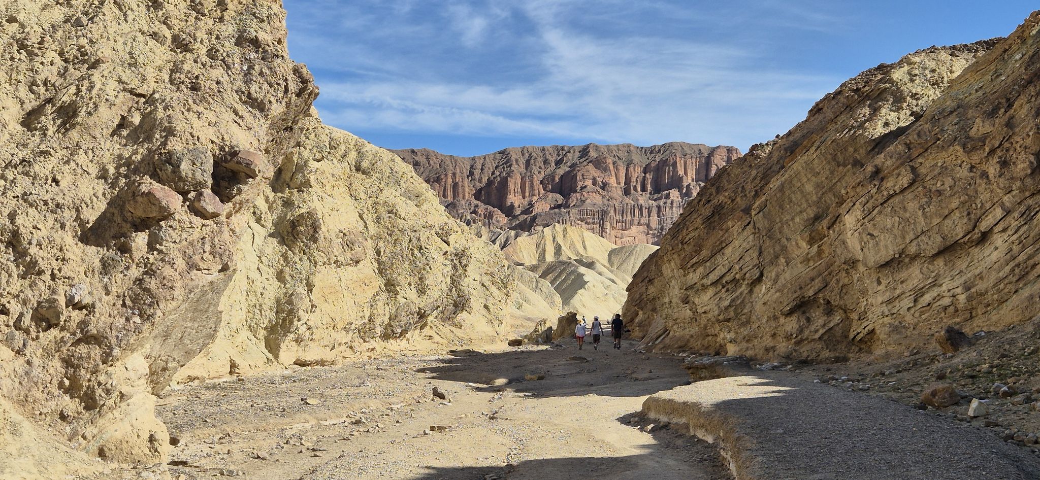 Hikers walk through a wide desert canyon with towering rock walls under blue sky. - Golden Canyon and Gower Gulch Loop Trail