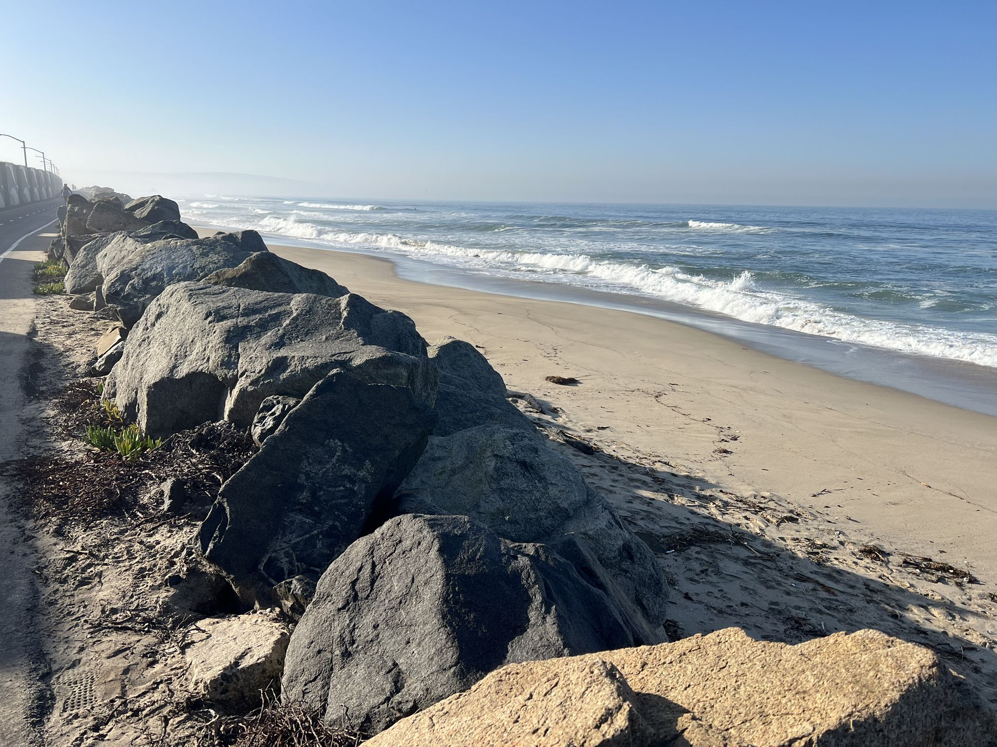 A rocky shoreline next to a sandy beach with ocean waves under a clear sky. - Inglewood, Watts, & Compton loop