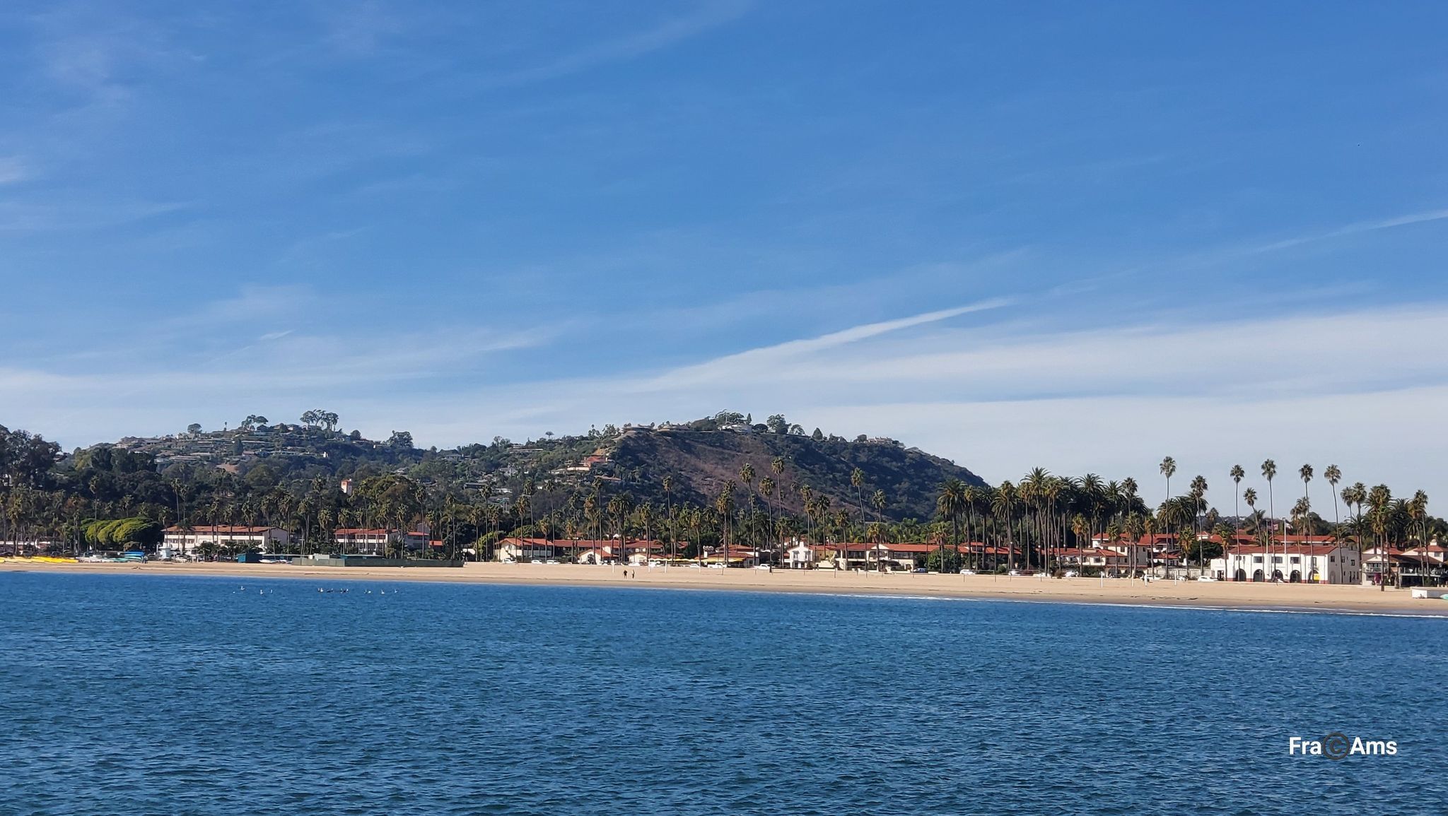 Blue ocean, sandy beach, coastal buildings, and green hills under clear sky. - Santa Barbara Stearns Wharf