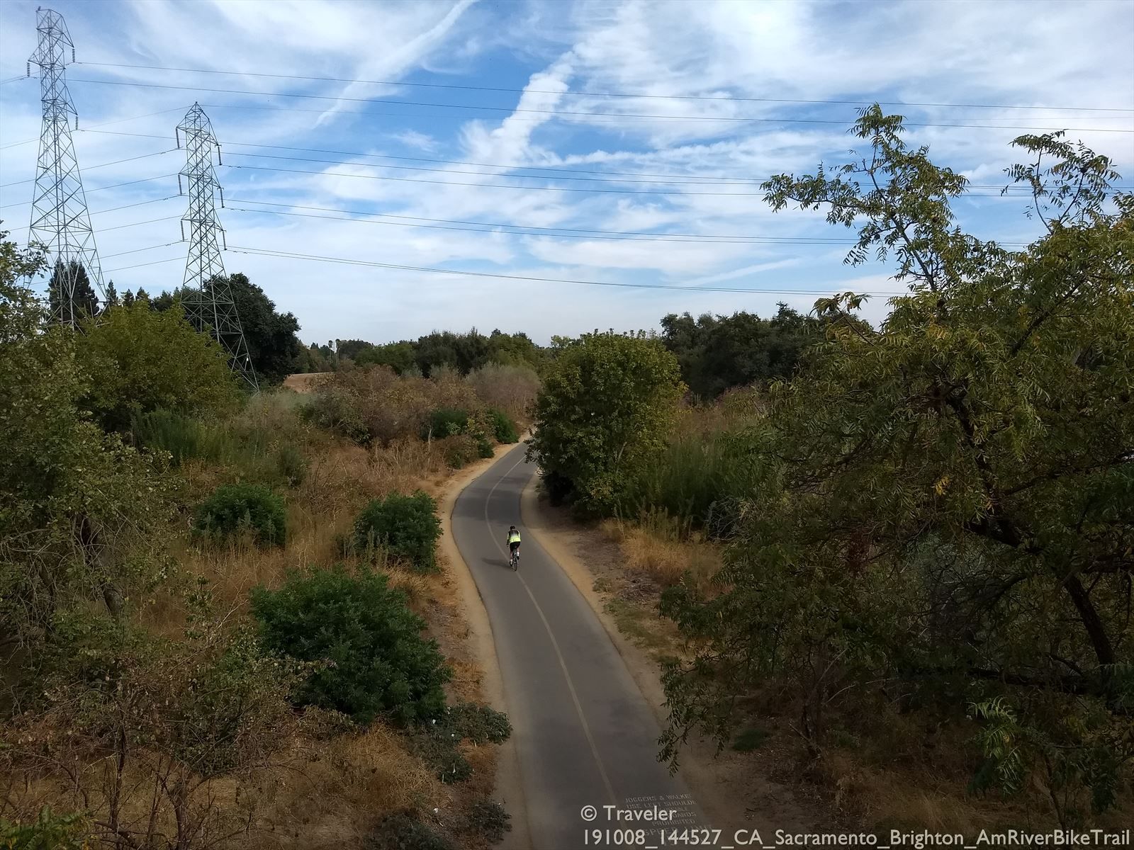 A person cycles on a winding paved path through green and dry trees. - Guy West Bridge – American River Bike Trail loop from Sacramento Valley Station