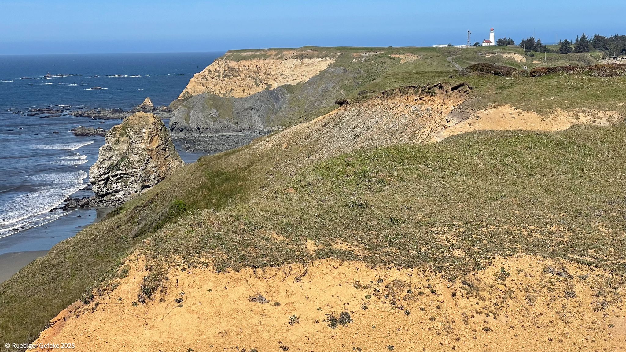 Sunny view of grassy coastal cliffs, rocky beach, and distant lighthouse. - Lighthouse Trail and Beach Trail