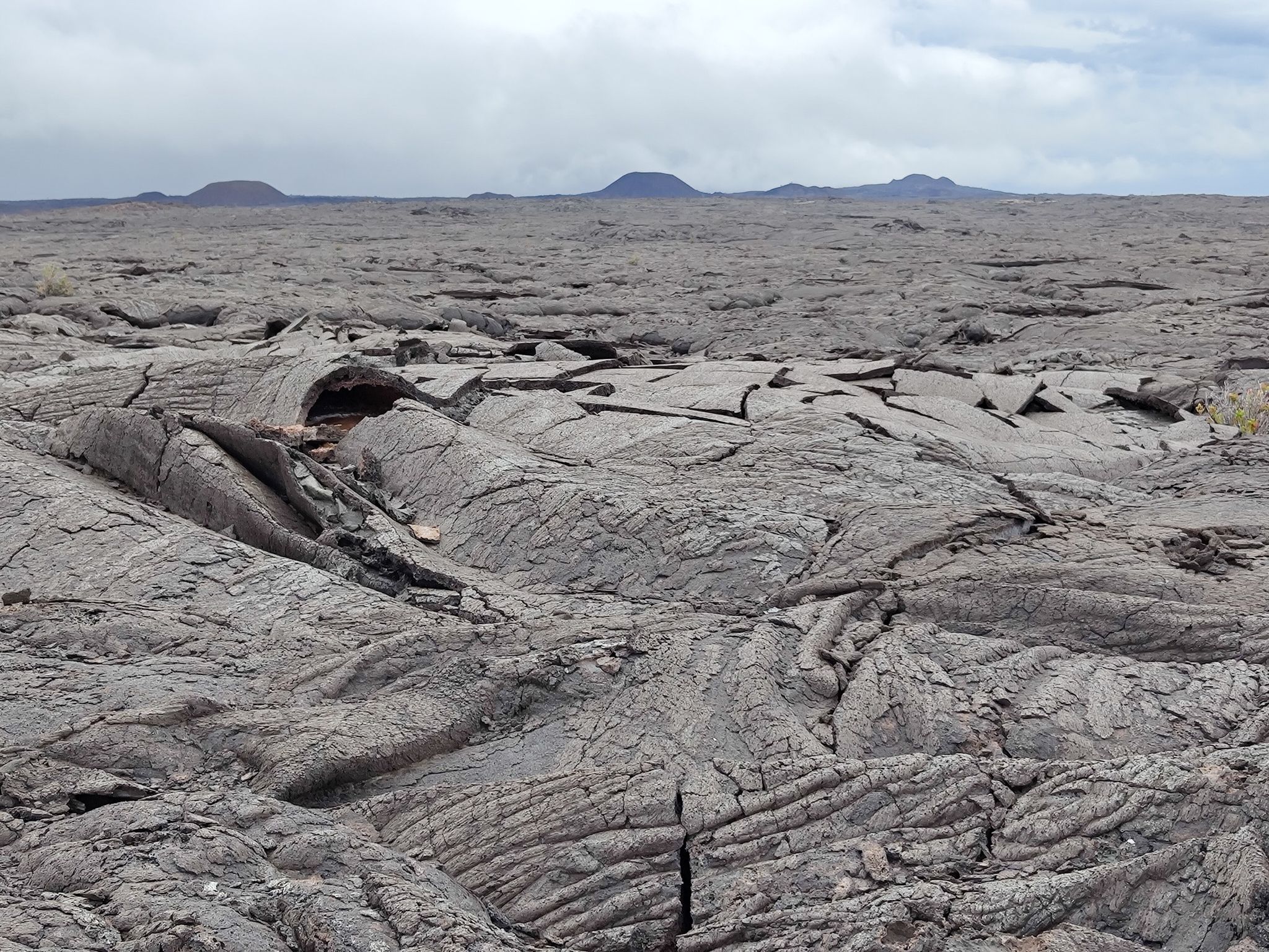 Extensive dark, cracked lava field under an overcast sky with distant hills. - Ka‘ū Desert Trail to Kamakai‘a Hills