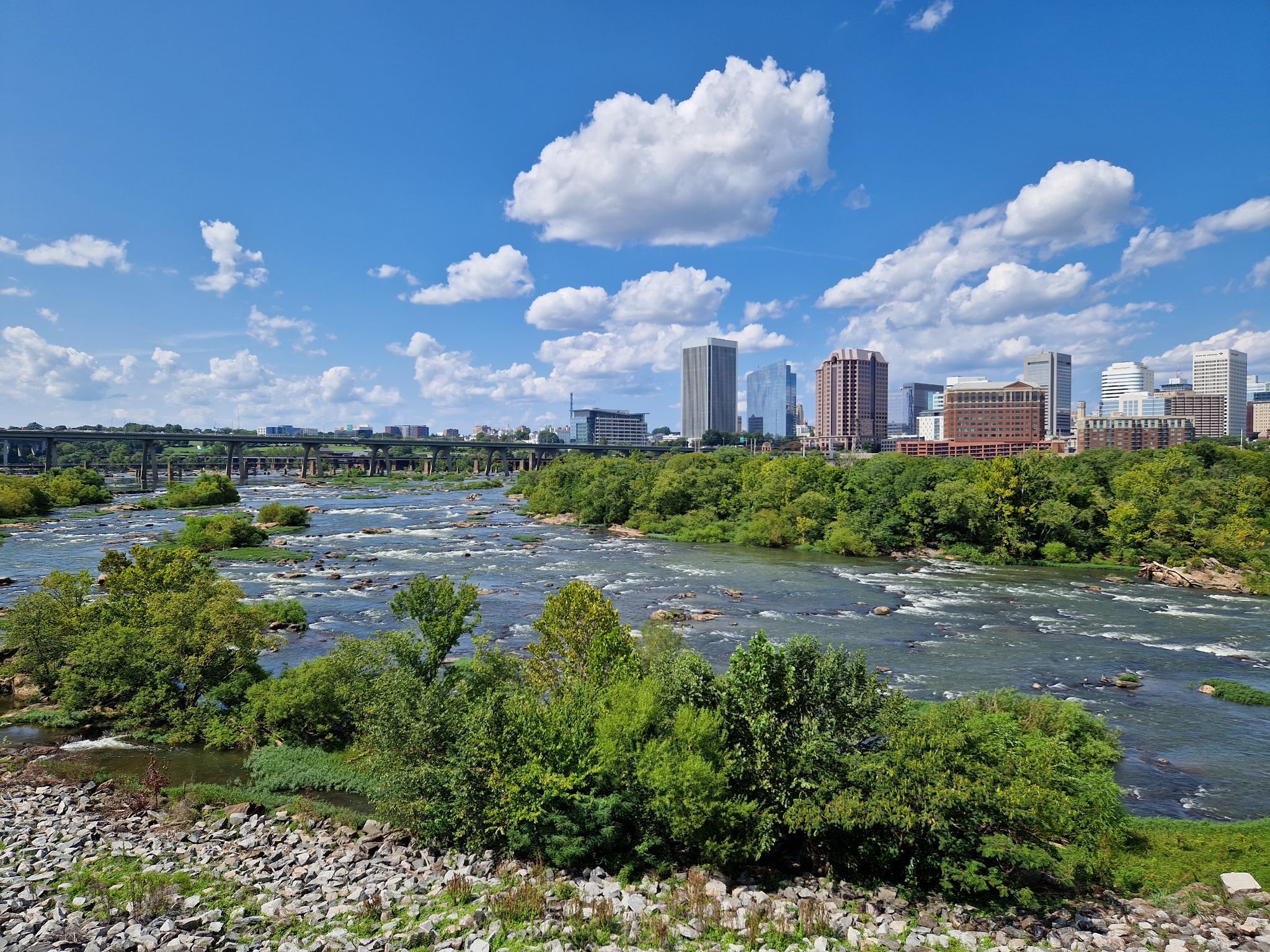 Rocky river with rapids and green trees, leading to a distant city skyline. - Manchester Floodwall and Slave Trail