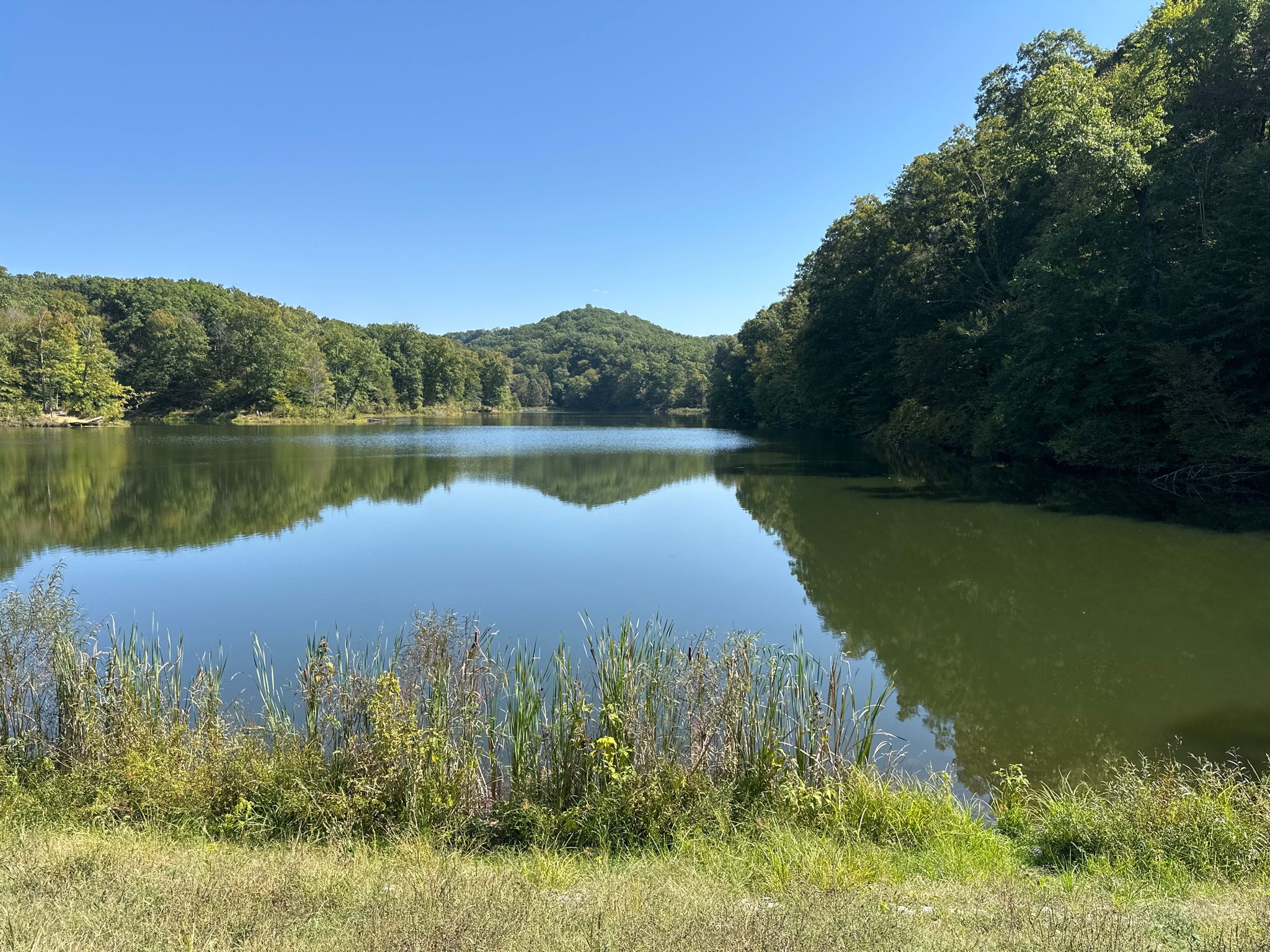 A calm lake reflects green hills and trees under a bright blue sky. - Ogle Lake loop from Brown County