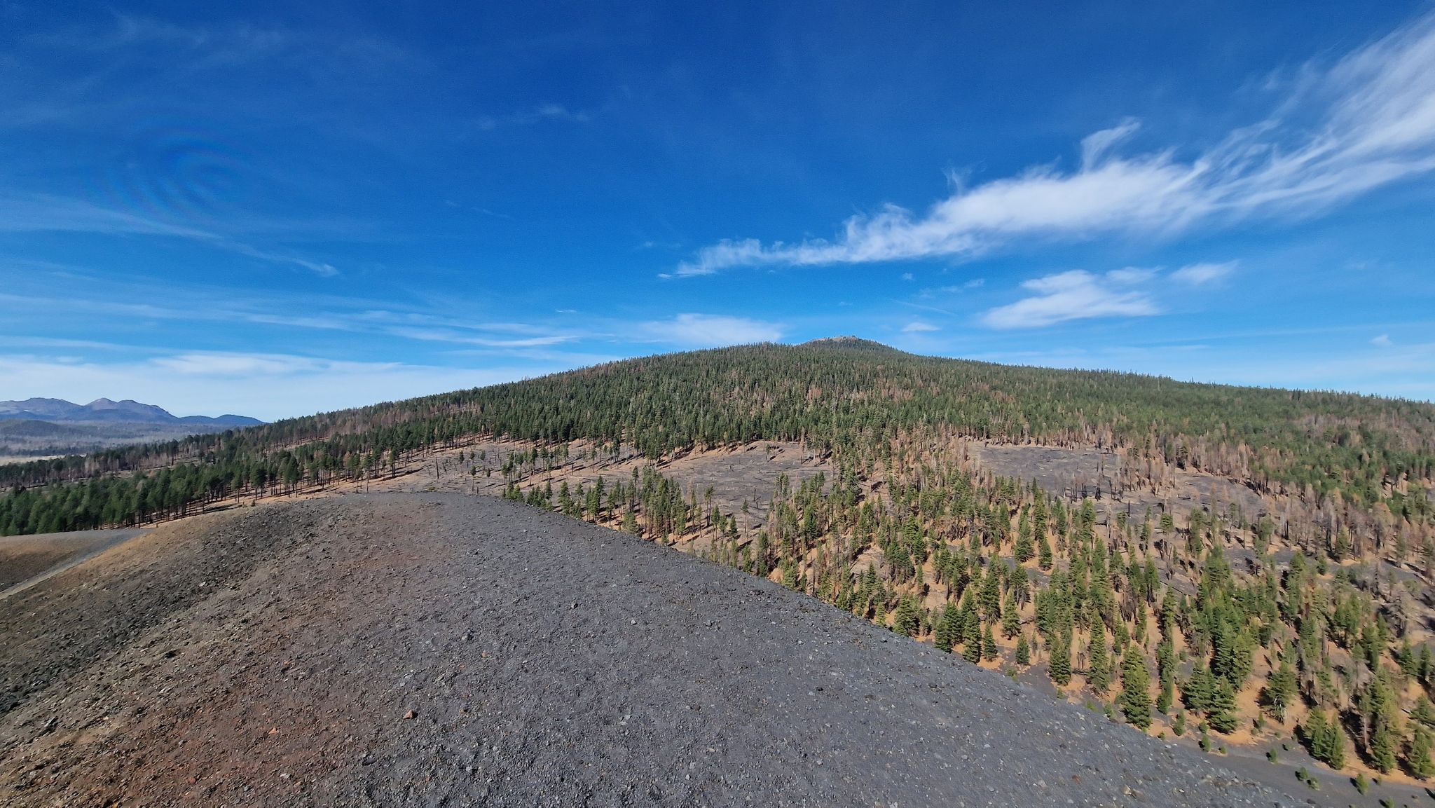 Dark rocky ridge leads to a forested hill under a bright blue sky. - Butte Lake, Snag Lake and Cinder Cone Loop