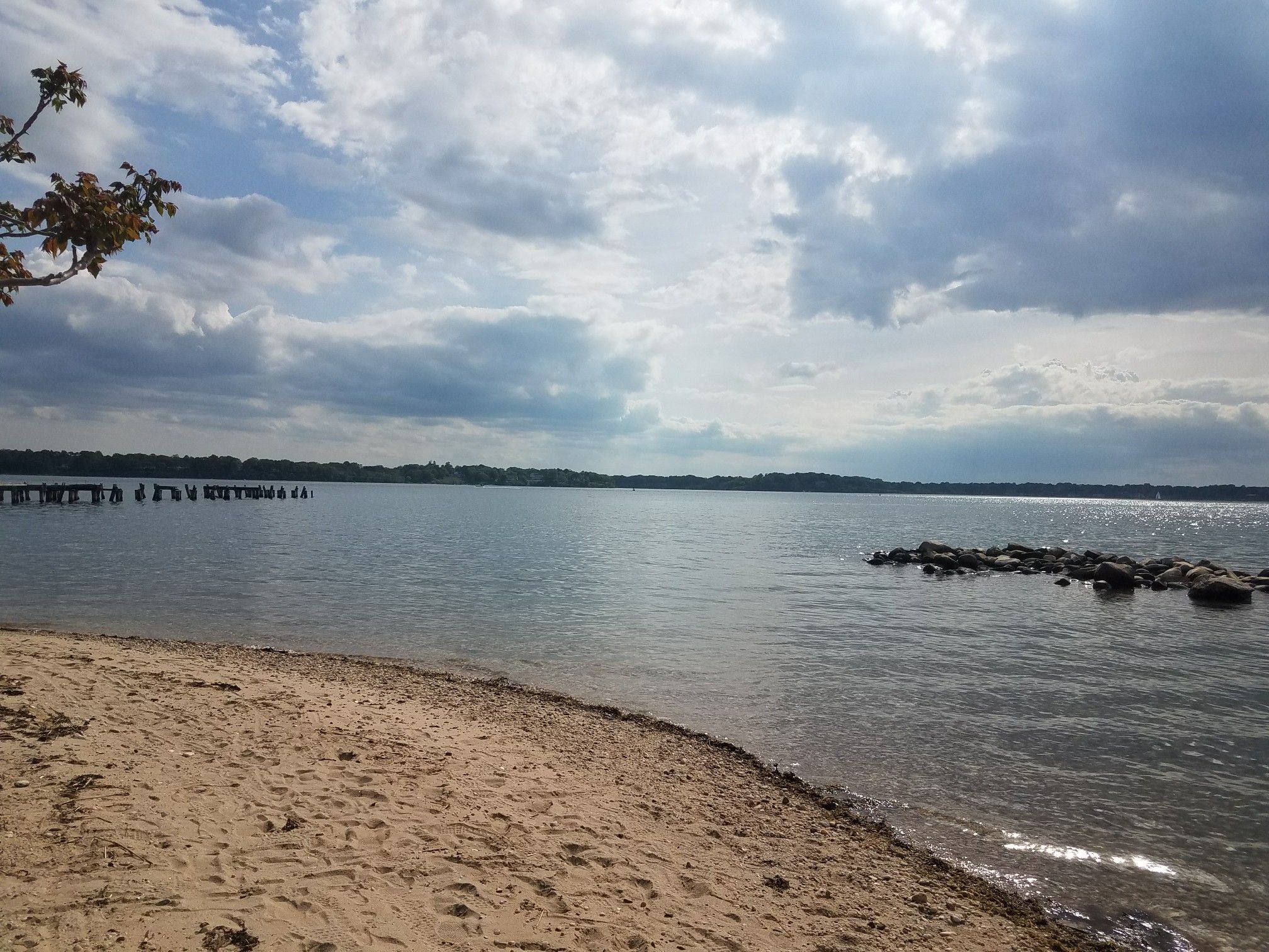 Sandy beach with calm water under a cloudy sky, distant pier structures. - McAllister County Park Loop