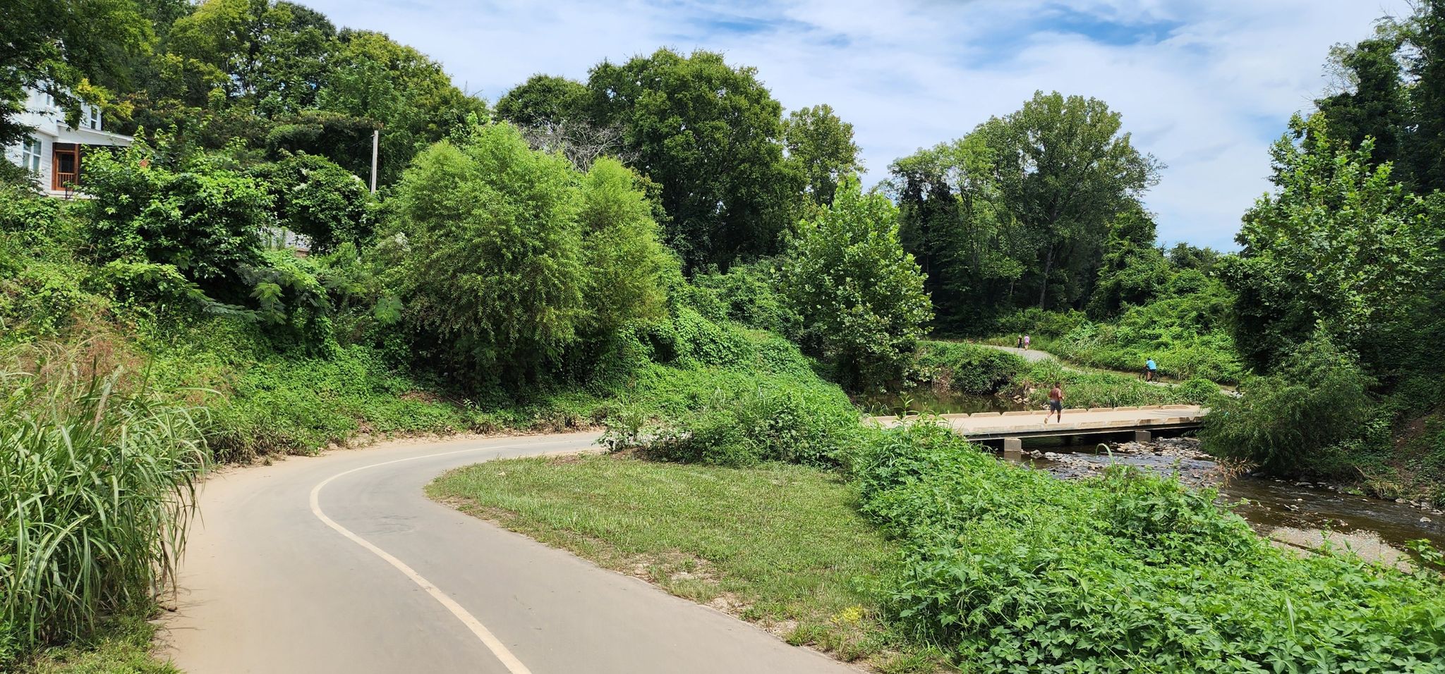 A winding paved path with lush green foliage, a person on a distant bridge. - Little Sugar Creek Greenway