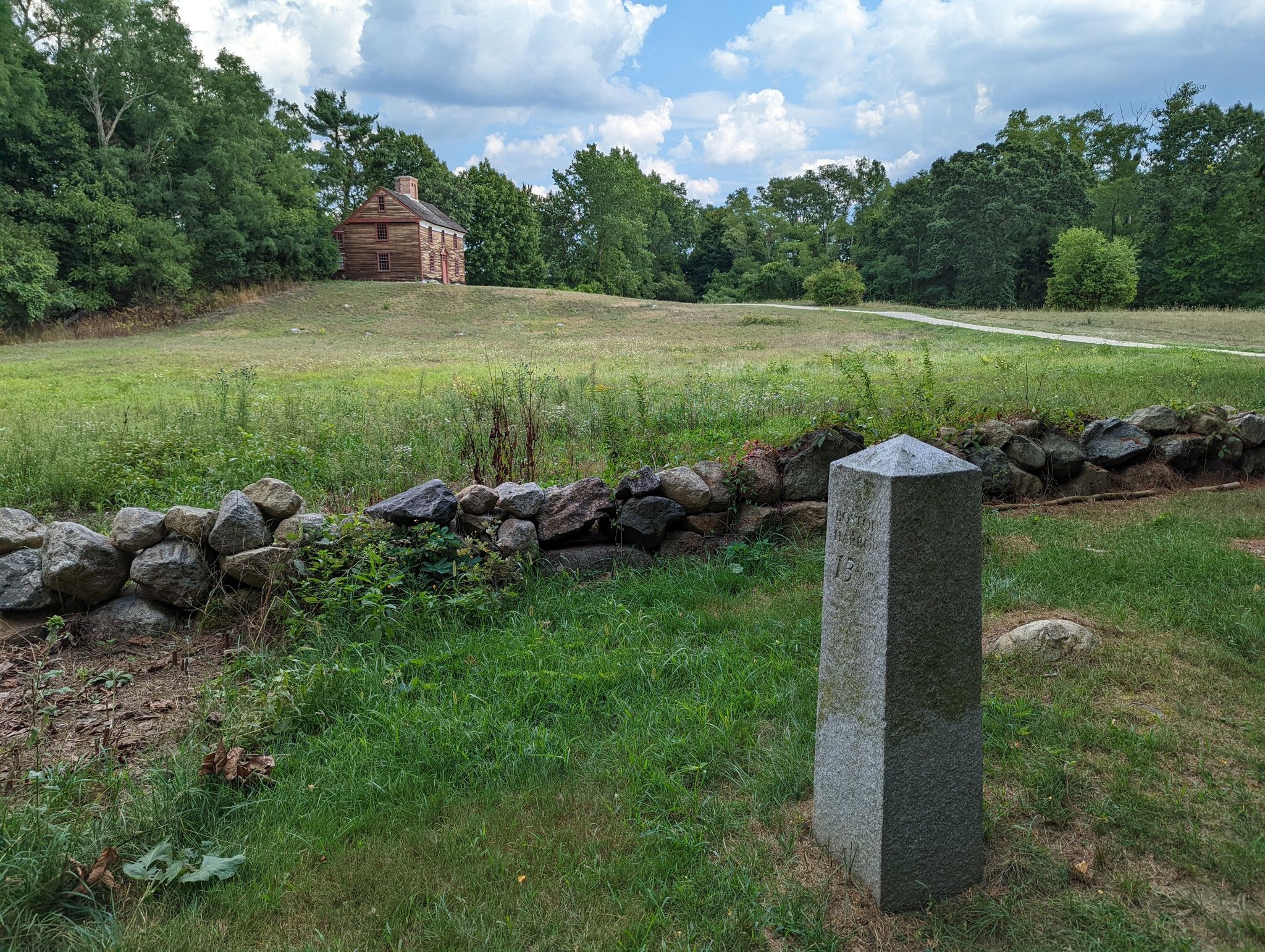 A stone marker and wall in a grassy field with a distant brick house. - Battle Road Trail – Battle Road Trail loop from Minute Man National Historical Park