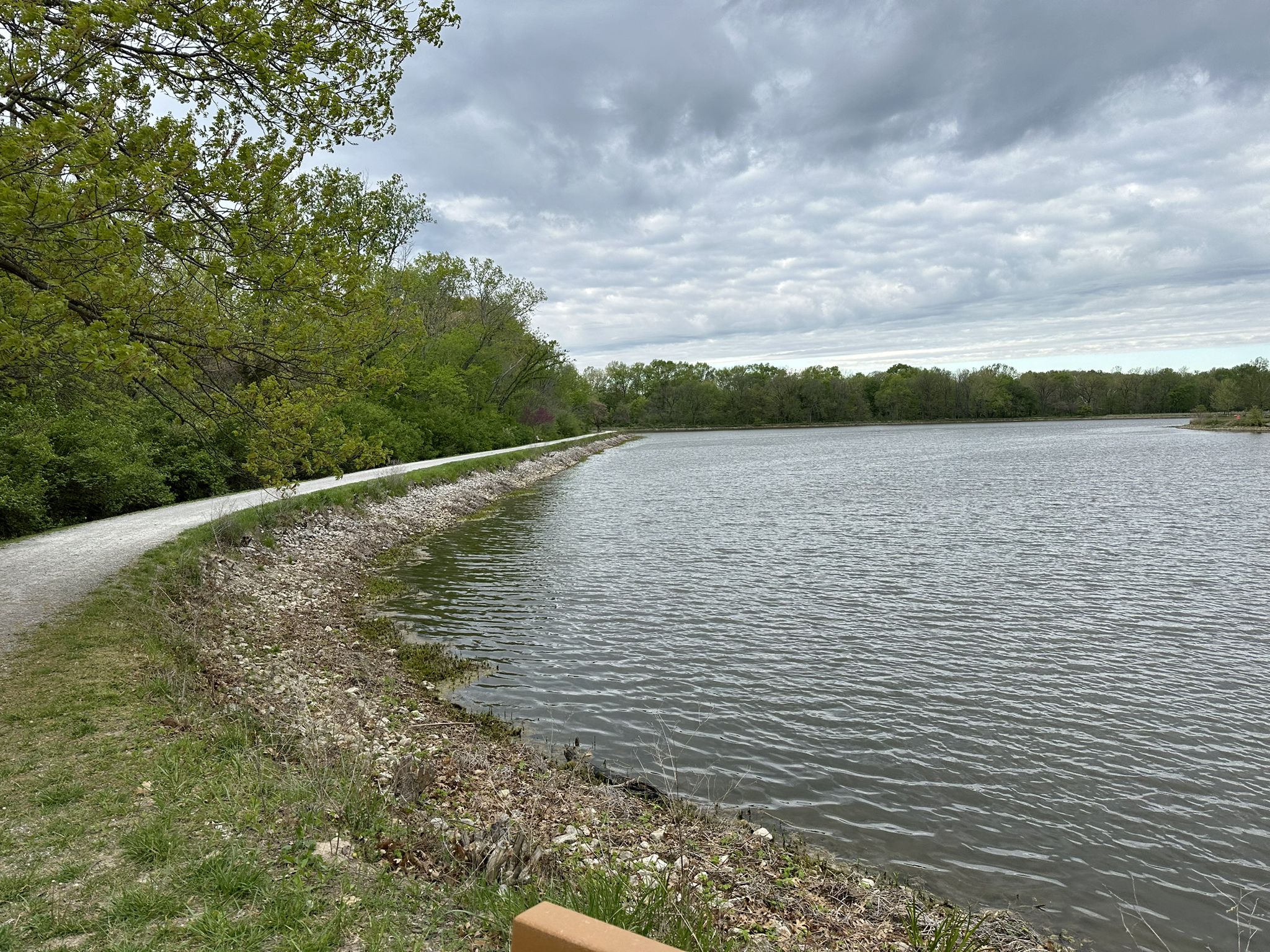 A gravel path curves along a lake under a cloudy sky, surrounded by green trees. - MKT Trail to Twin Lakes Loop