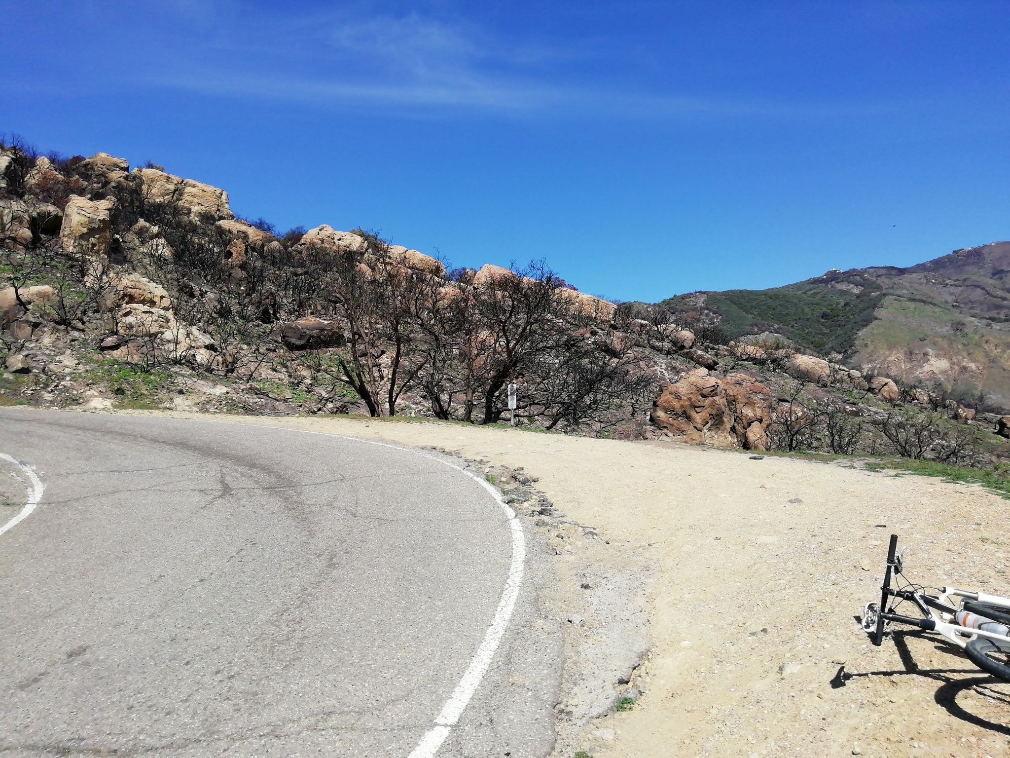 A curved road and dirt shoulder with a bicycle, rocky burned hills. - La Cumbre Peak loop