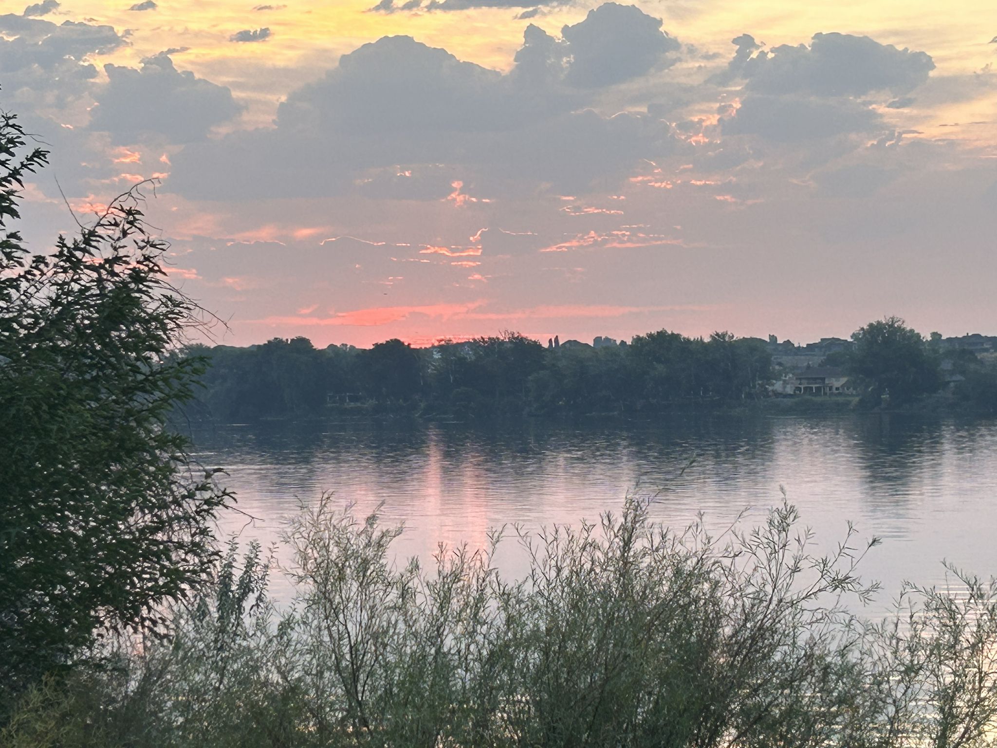A colorful sunset over a river, with trees and bushes in the foreground. - Columbia Point Marina Park – Riverfront Trail loop from Bradley at River's Edge