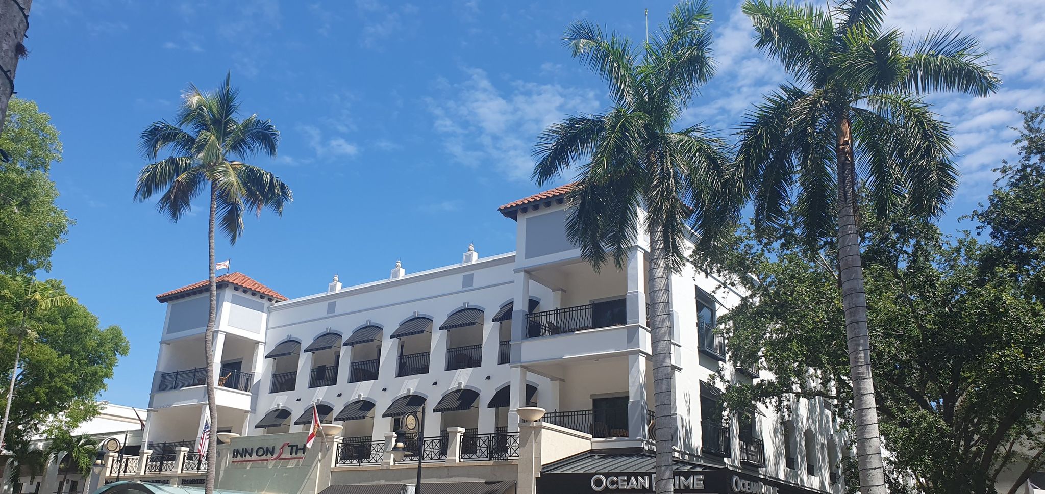 A white building with palm trees under a bright blue sky. - Sugden Plaza – 5th Avenue South (Old Naples) loop from Naples