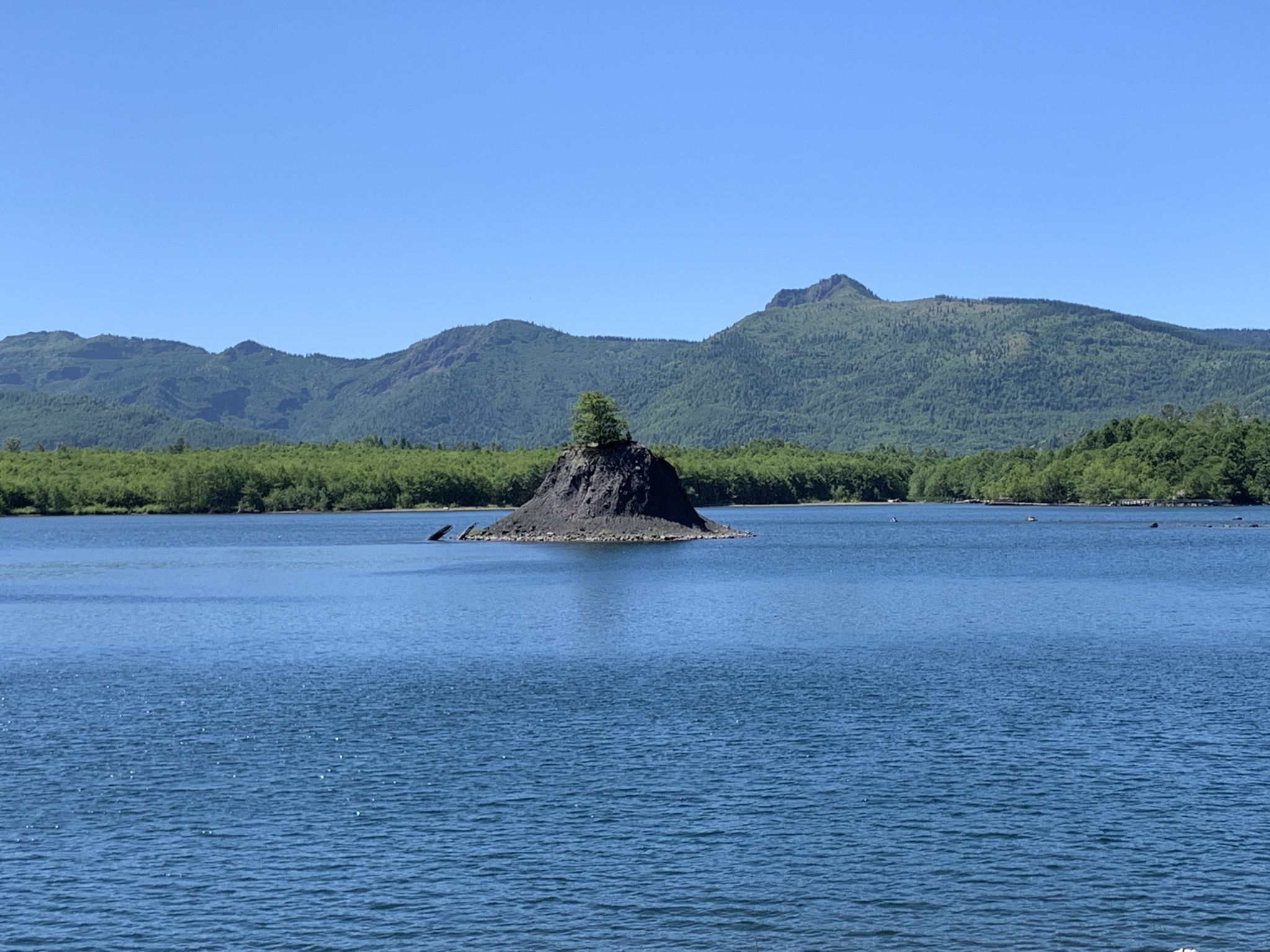Small tree-covered island in a wide blue lake with distant green mountains. - Lakes Trail