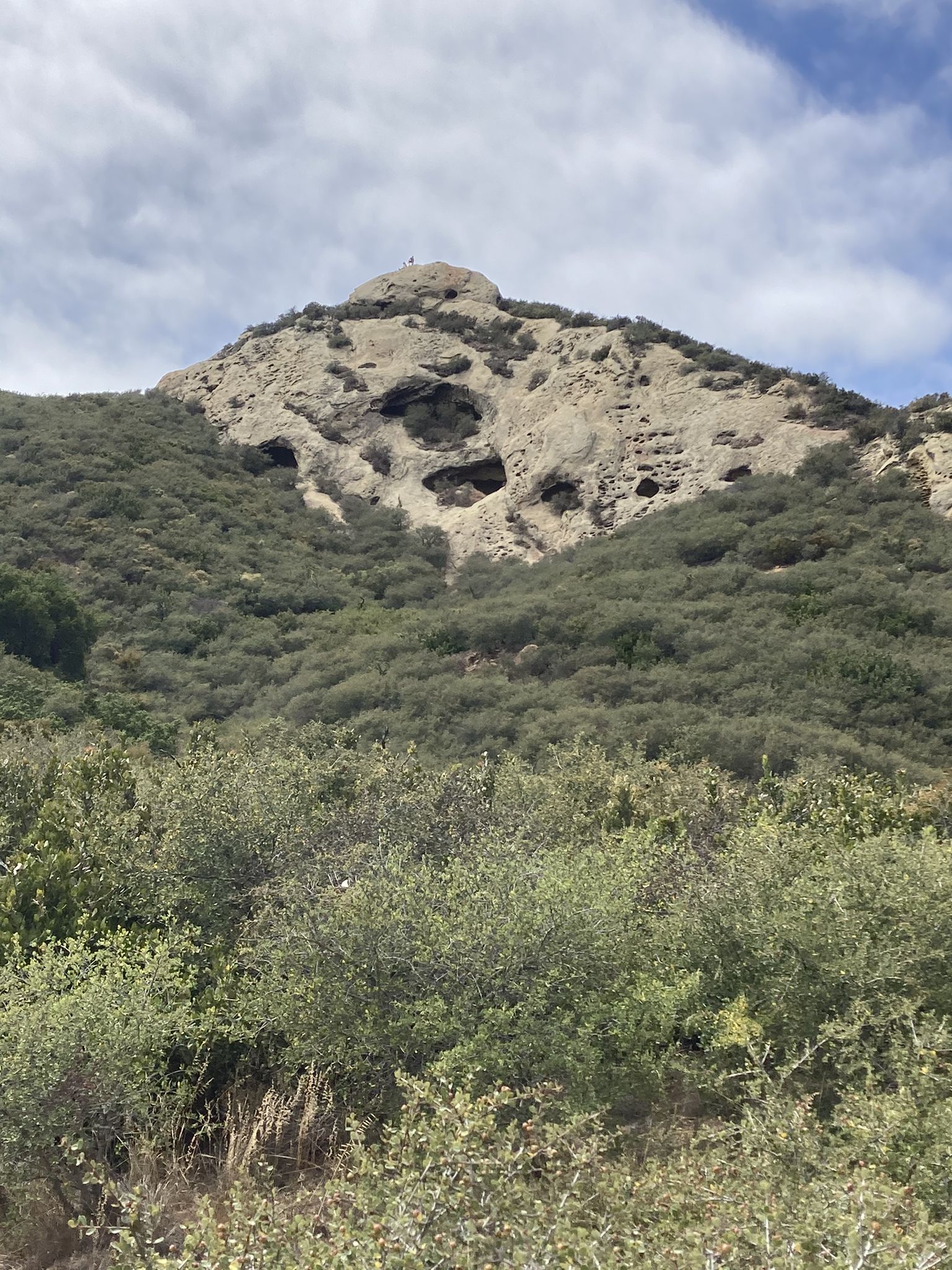 Rocky mountain with dark holes, green shrubs, and small figures on peak. - Gaviota Wind Caves run via Beach to Backcountry Trail