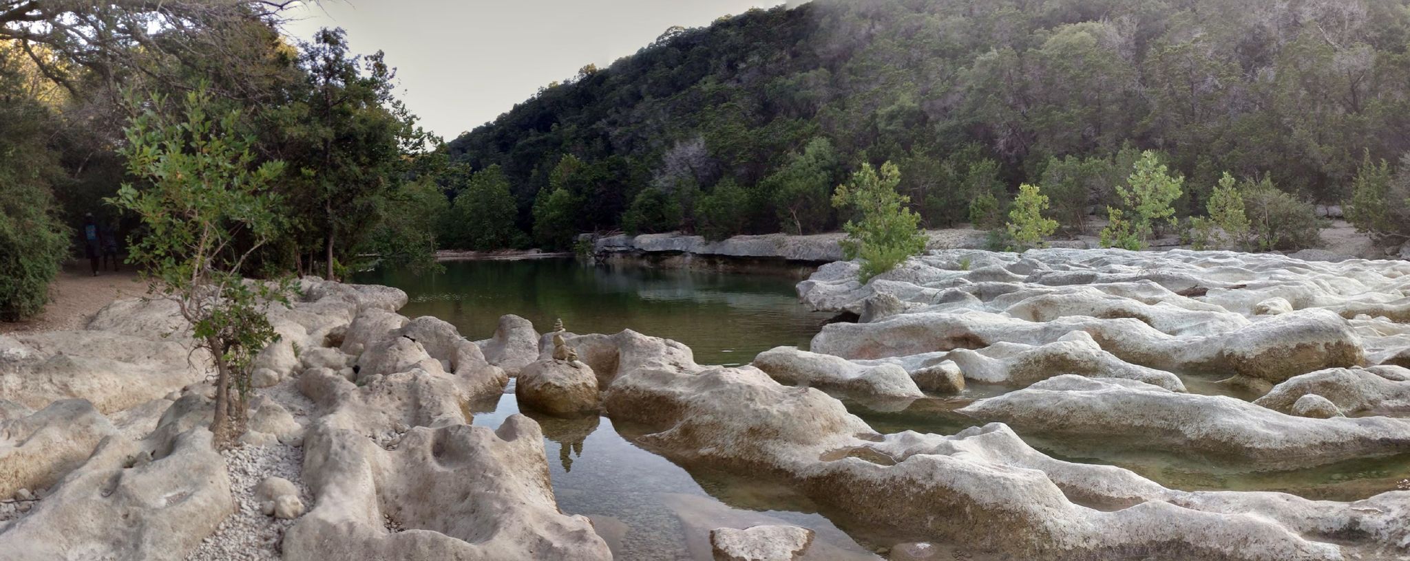 Panoramic view of a calm river with rocky banks and forested hills under a bright sky. - Barton Creek Sculpture Falls loop