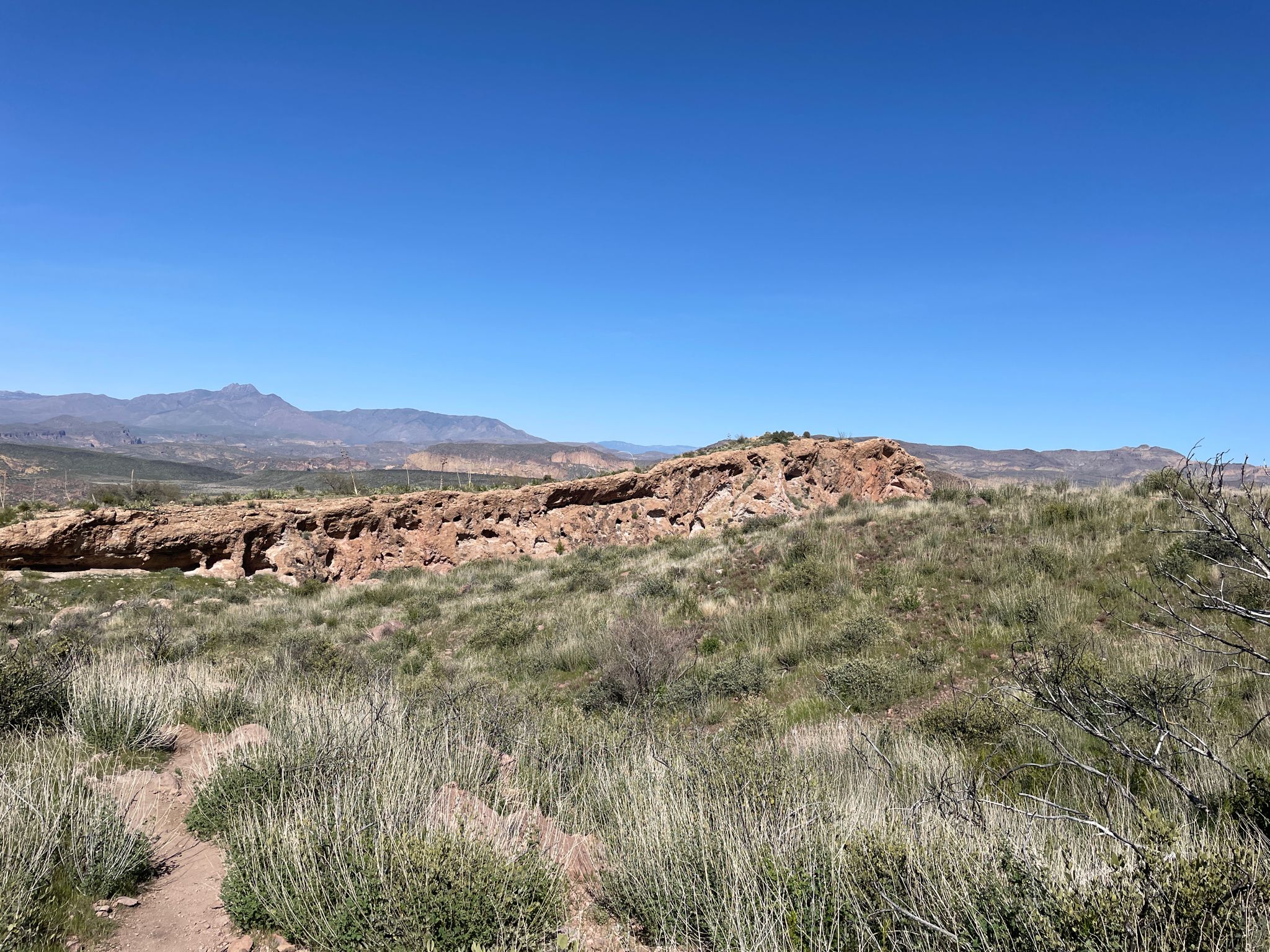 Sunny desert landscape with a rocky ridge, green brush, and a dirt path. - Massacre Falls Trail