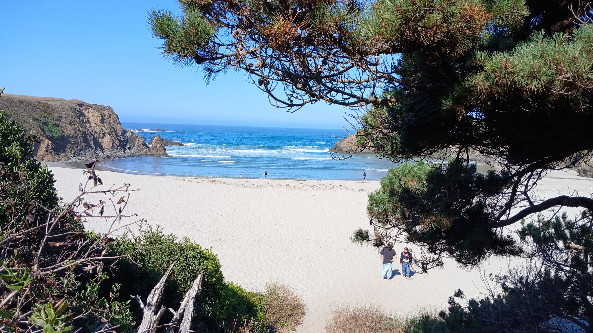 Sunny beach with blue ocean, rocky cliffs, and people walking on sand. - Ecological Staircase Trail