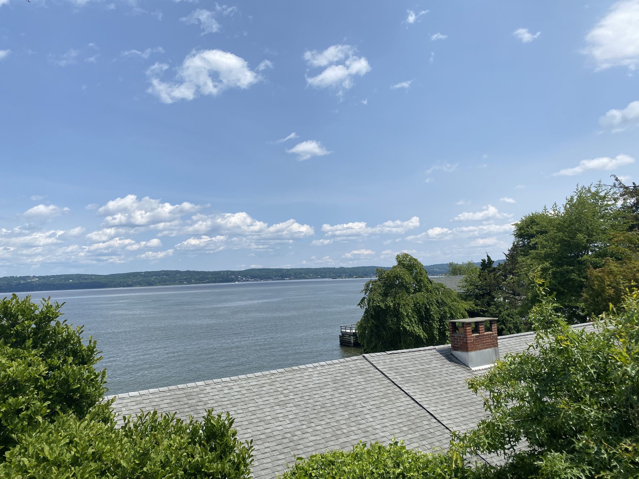Expansive lake view over a house roof and green trees on a bright, cloudy day. - Lake DeForest Loop
