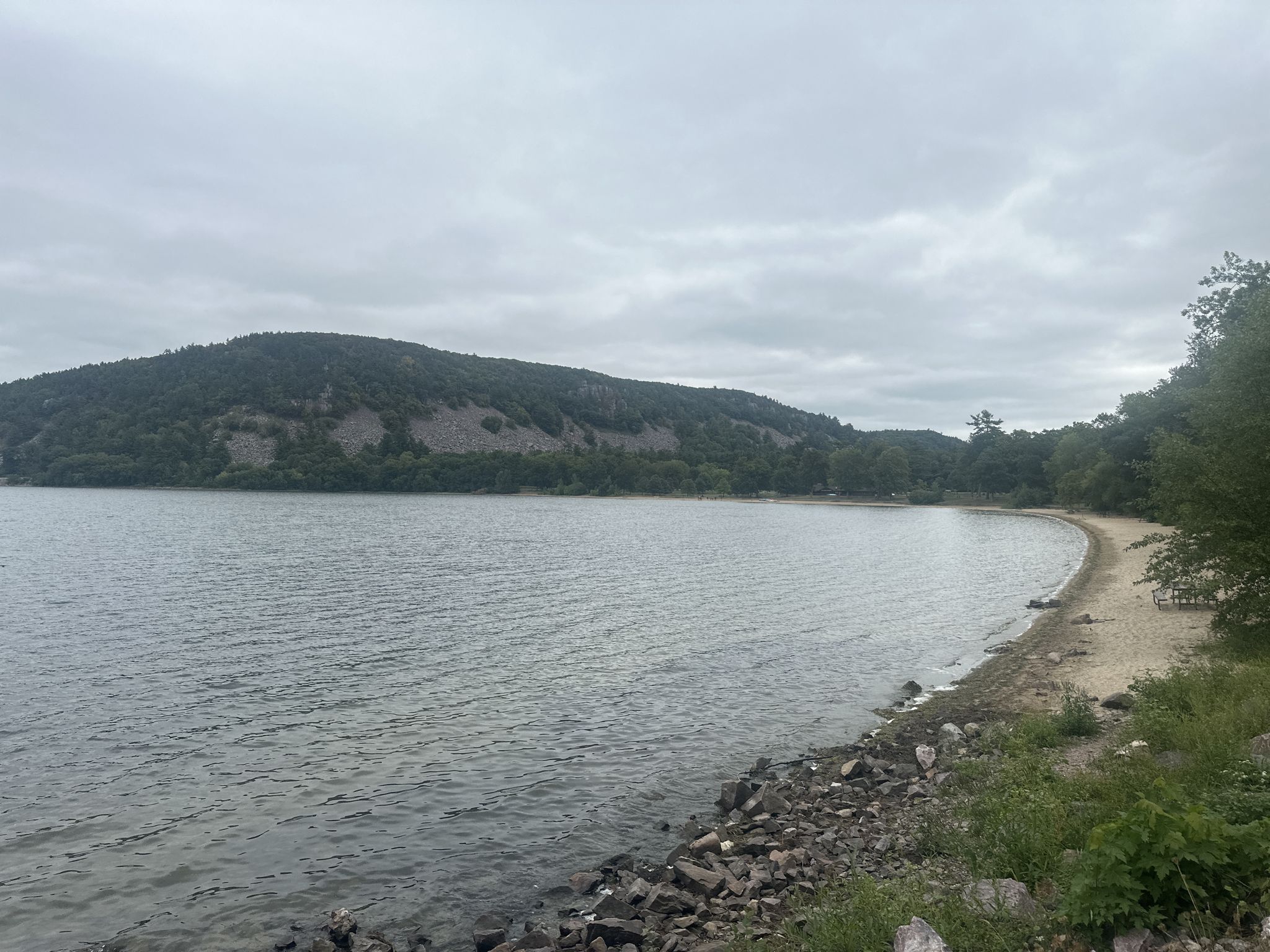 A calm lake with a rocky shore and forested hills under an overcast sky. - Devil's Lake State Park – Devil's Lake State Park loop from Baraboo