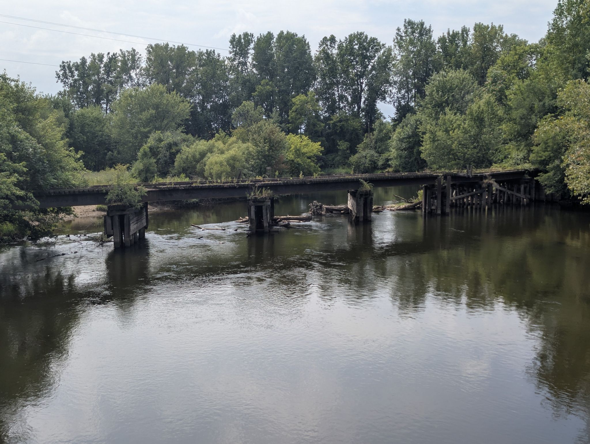 Old, dilapidated bridge over a calm river, surrounded by green trees. - Kalamazoo River Valley Trail