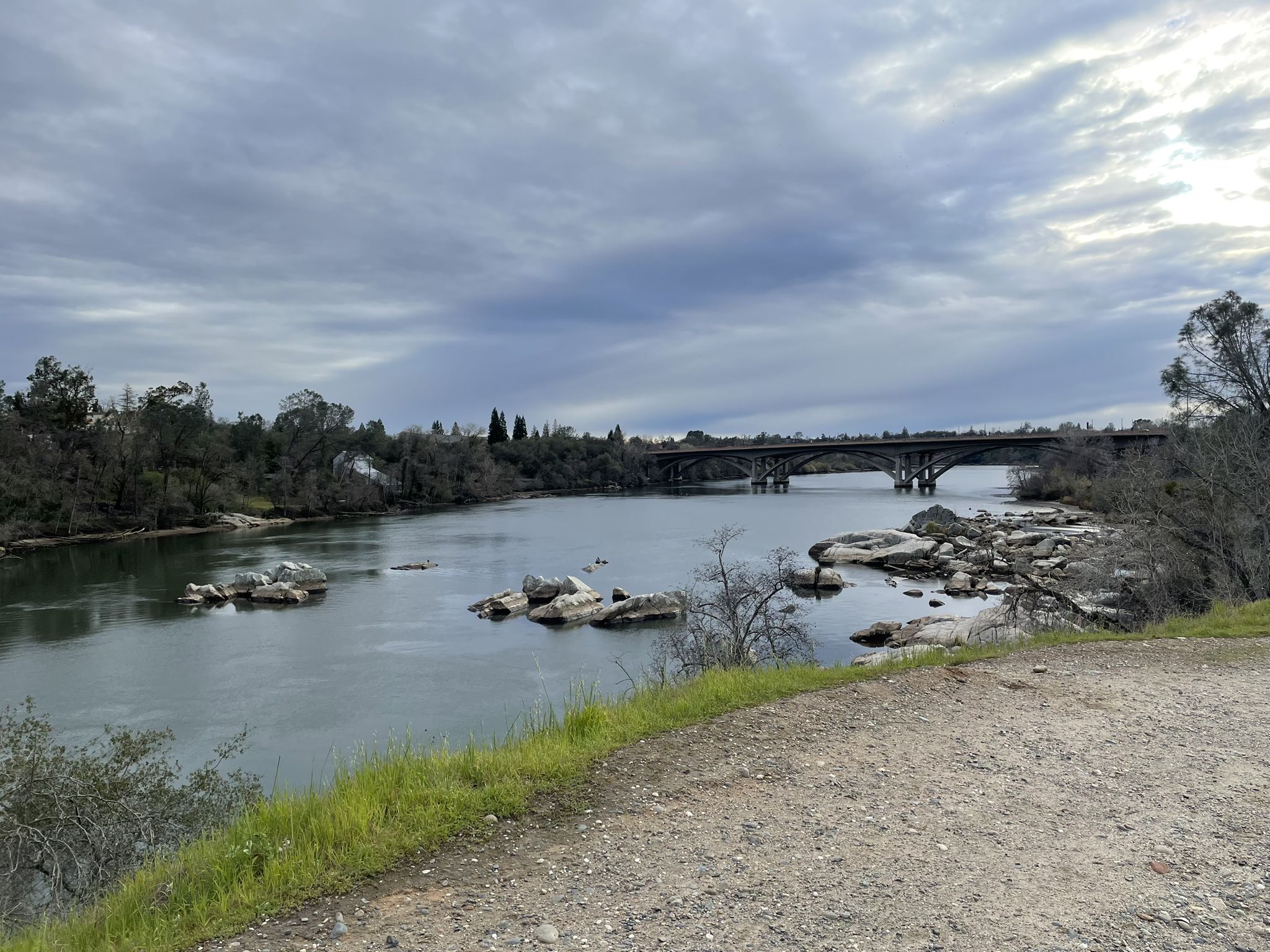 Overcast day overlooking a wide river with rocks and a distant bridge. - Folsom Historic Truss Bridge – Folsom Historic Truss Bridge loop from Folsom