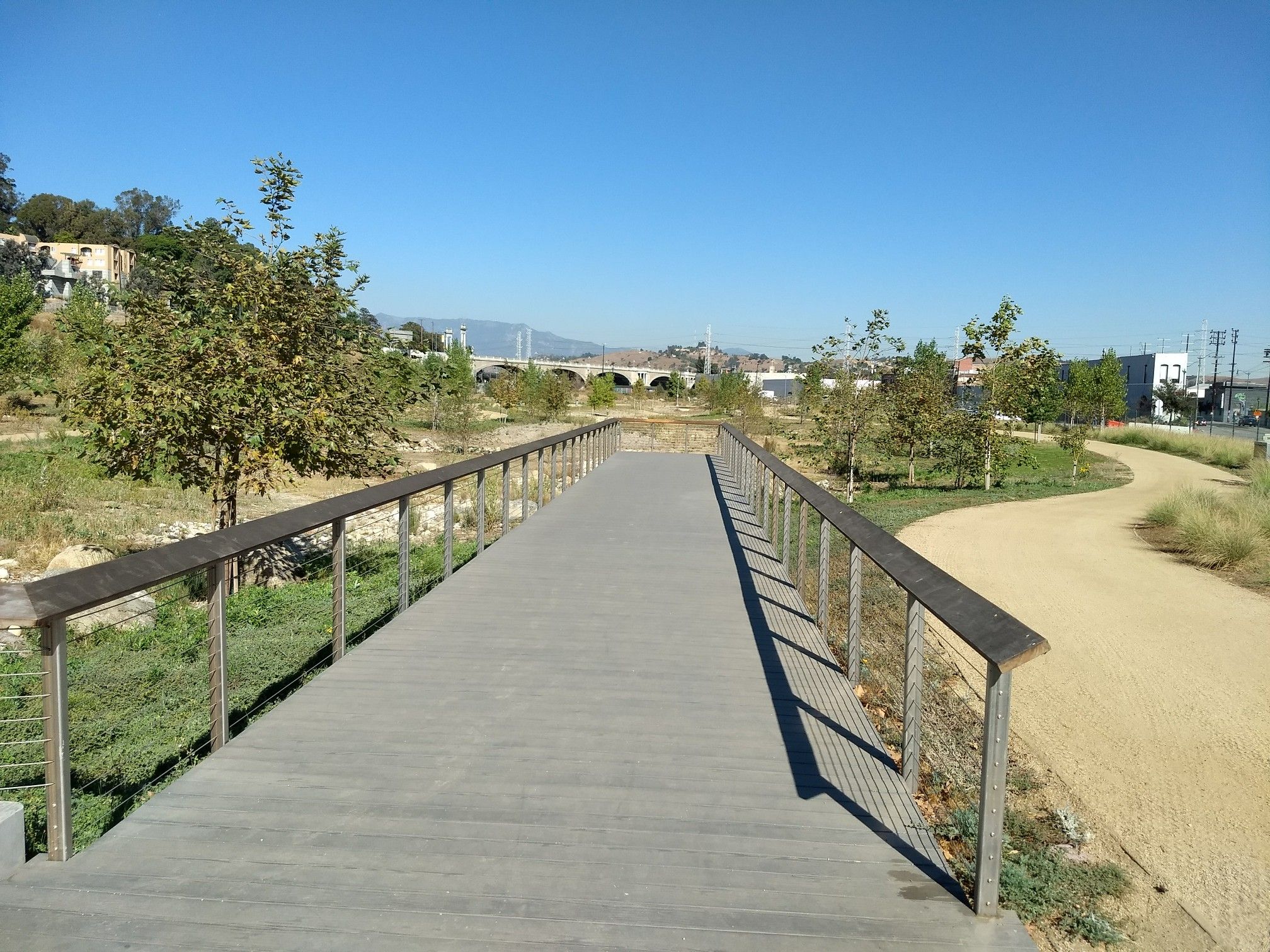 A wooden pedestrian bridge extends over a grassy landscape under a clear blue sky. - Los Angeles State Historic Park Loop Trail