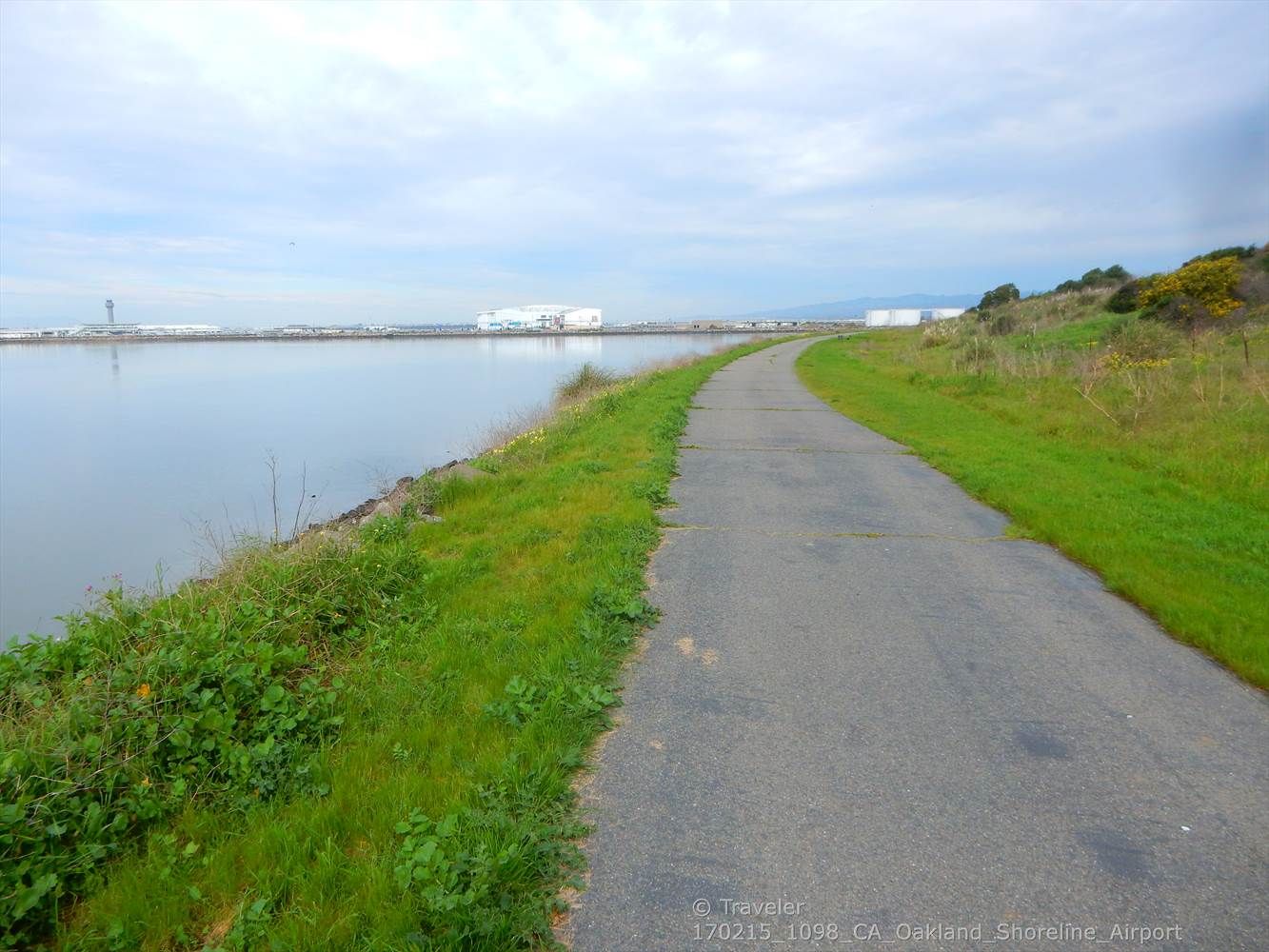A paved path runs beside calm water with distant industrial buildings under clouds. - San Francisco Bay Trail: Oyster Bay Loop