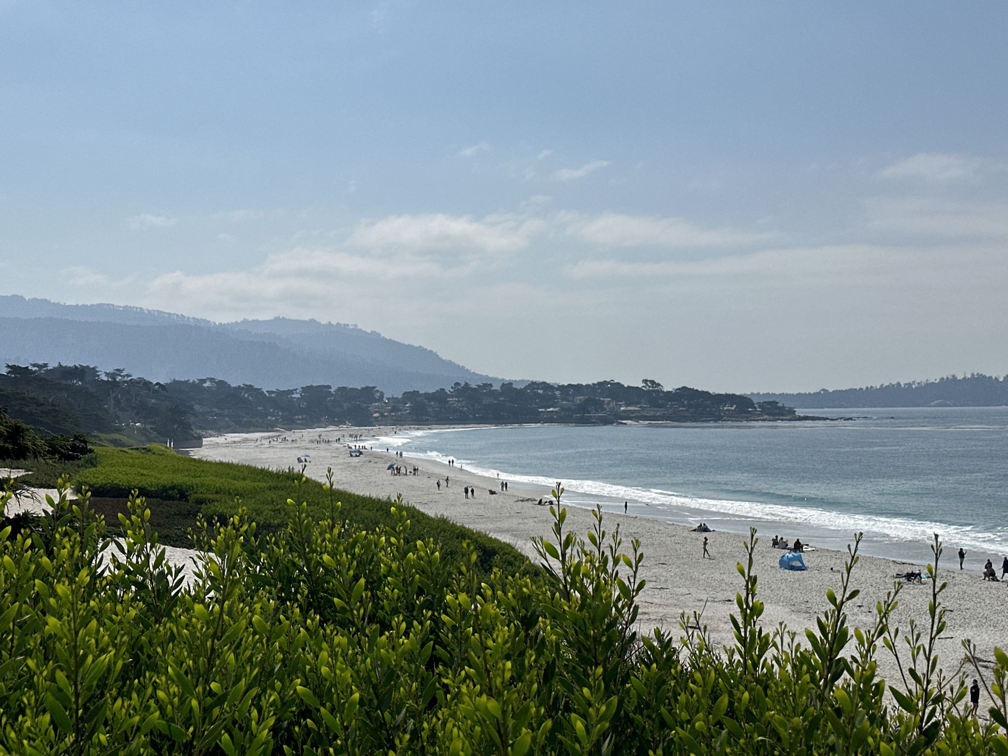 Wide sandy beach with ocean, distant mountains, and scattered people under bright sky. - Carmel Beach