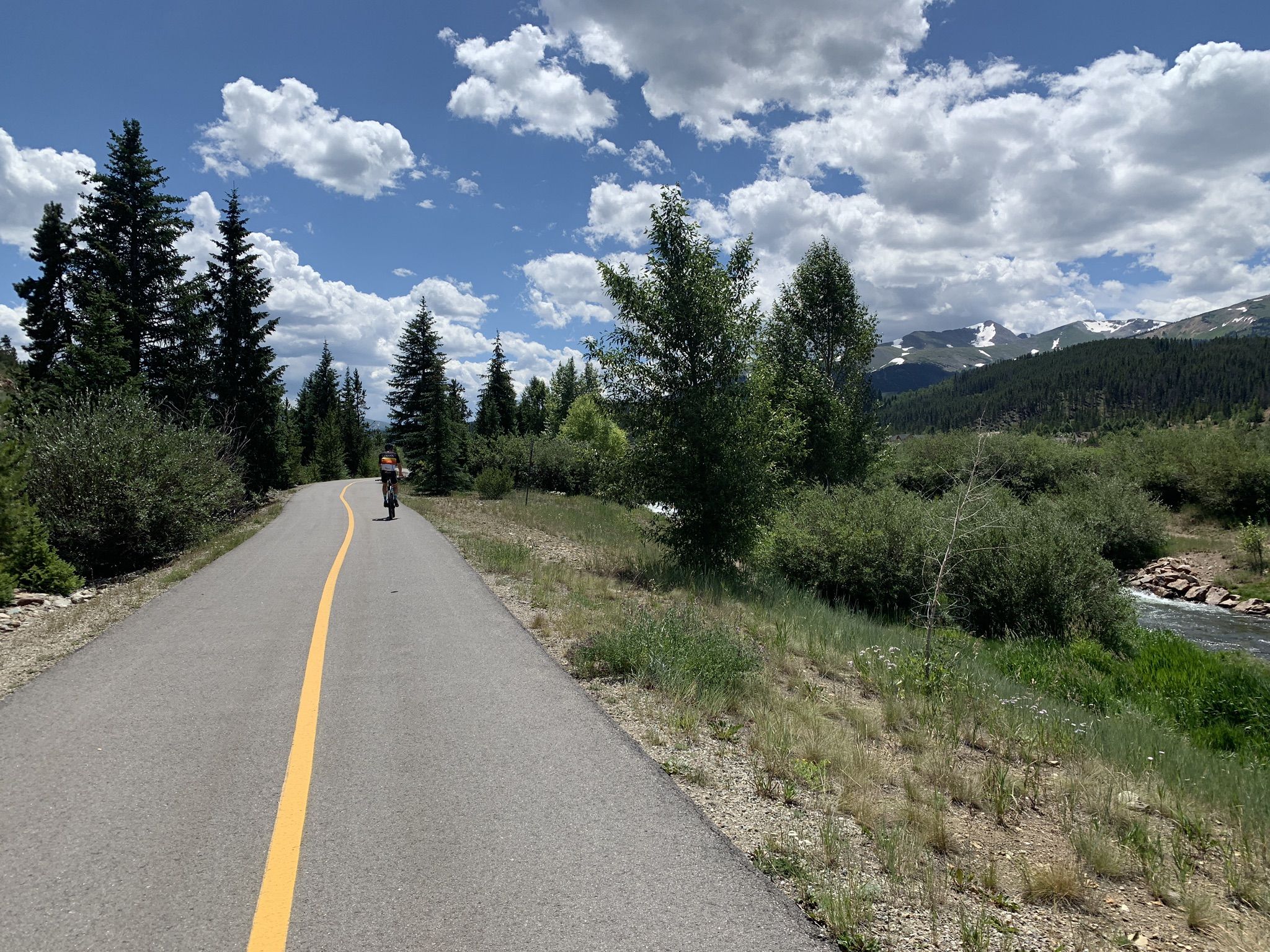 A person cycles on a paved path beside a river, with mountains and trees. - Upper Blue Recpath