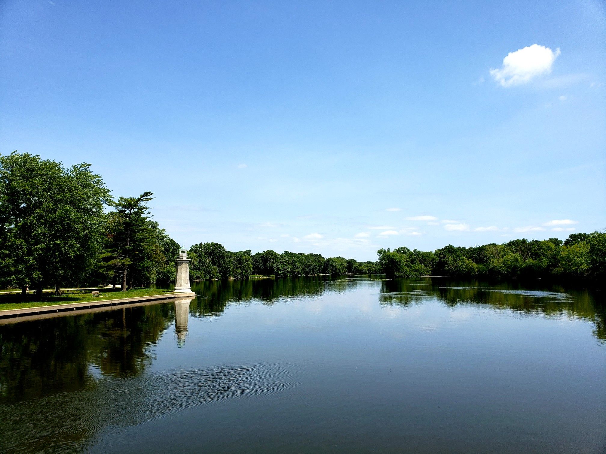 Wide river with a white lighthouse on the bank under blue sky. - Fabyan Forest Preserve – View of Fabyan Windmill loop from St. Charles