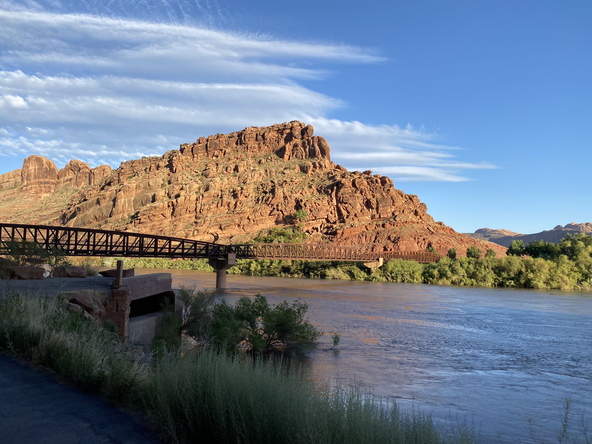 Metal bridge over a wide river, large red rock mountain, green trees, sunny. - Moab Canyon Pathway