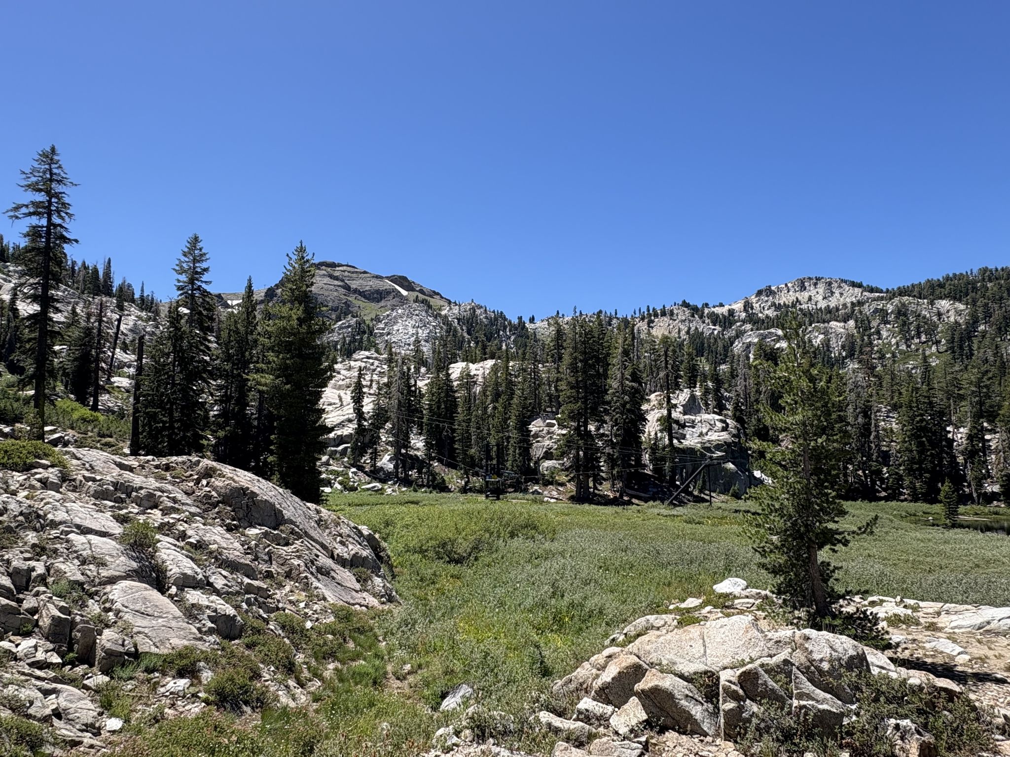 Sunny mountain landscape with rocky foreground, green meadow, and pine forest. - Squaw Valley – Squaw Valley village loop from Olympic Valley