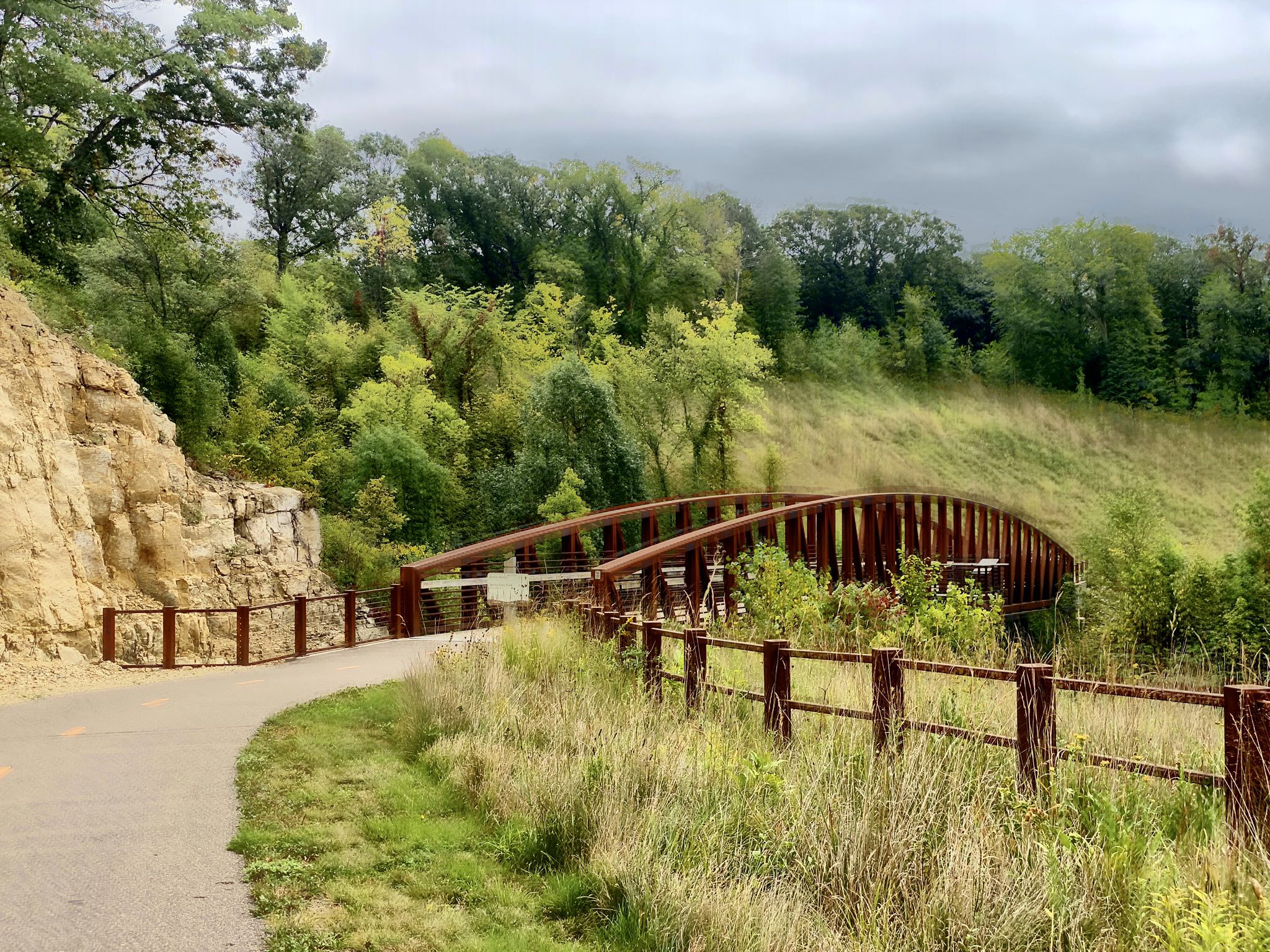 Curved wooden bridge on a paved path through green hills and trees. - Spring Lake Overlook – Spring Lake Park Reserve loop from Nininger Township