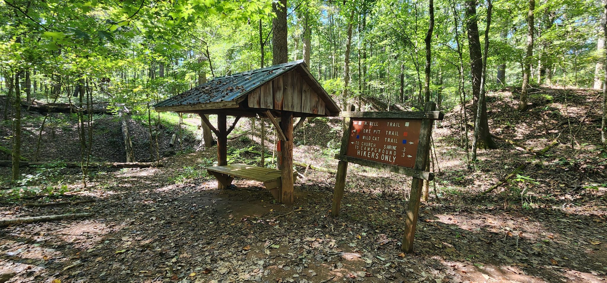 A rustic wooden shelter with a bench and sign in a sunny forest. - Ore Pit Loop Trail